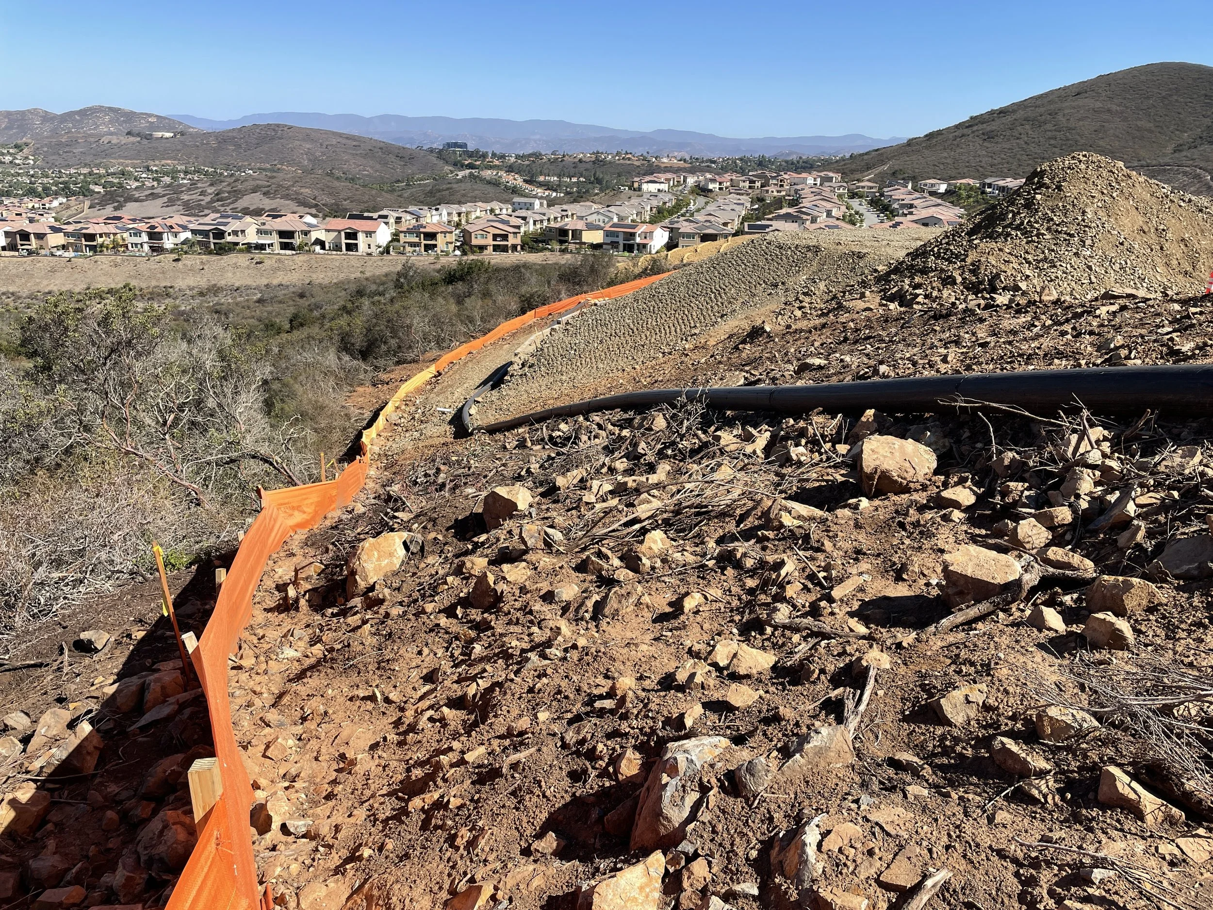 Construction site with rocky terrain, orange safety fencing, and view of residential neighborhood in the background.