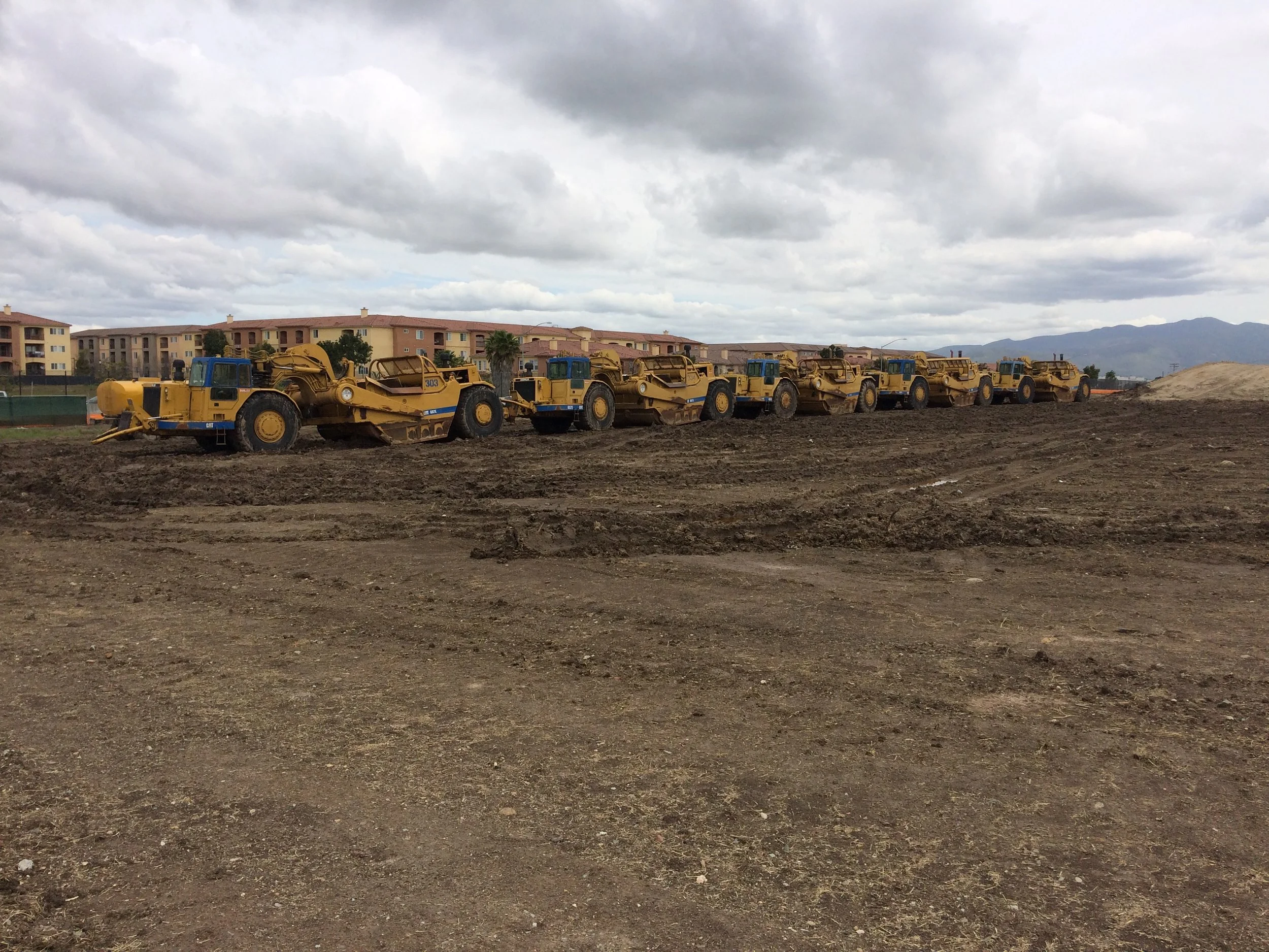 Row of yellow construction vehicles on a barren field with buildings in the background under a cloudy sky.