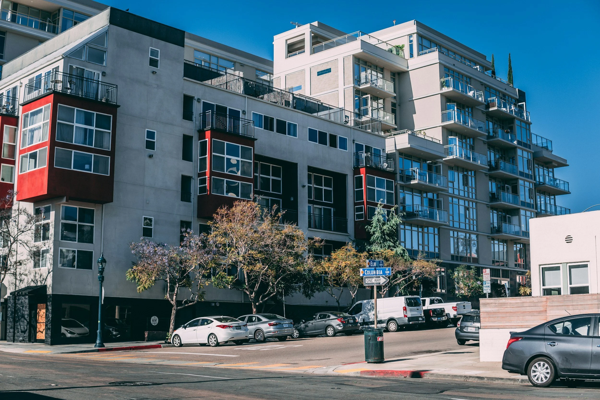 Modern urban apartment buildings with balconies and large windows, street view with parked cars and street signs.