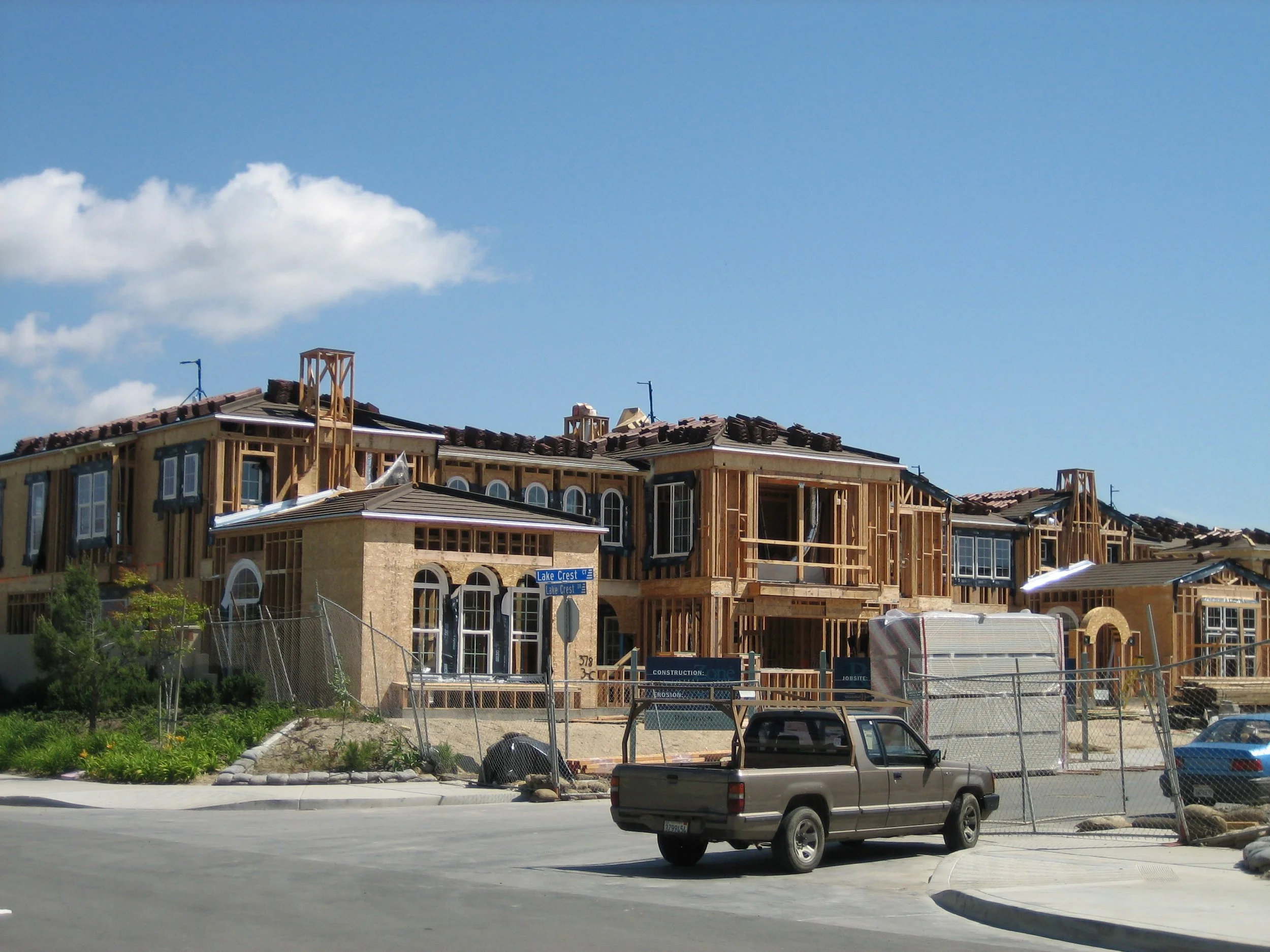 Residential houses under construction with wooden frames, surrounded by a chain-link fence and construction signs.