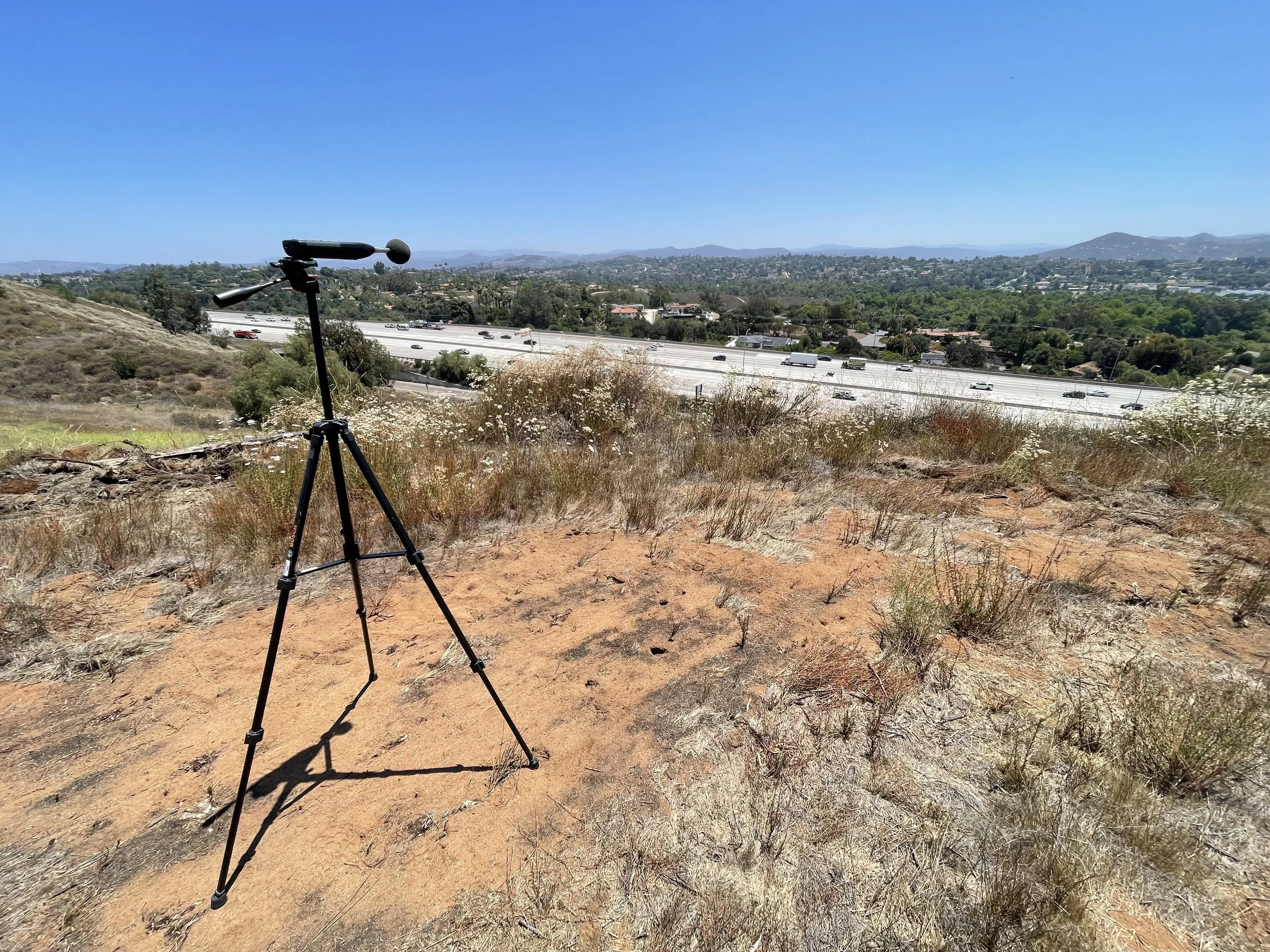 A camera tripod set up on dry, grassy terrain overlooking a highway and a cityscape in the distance under a clear blue sky.