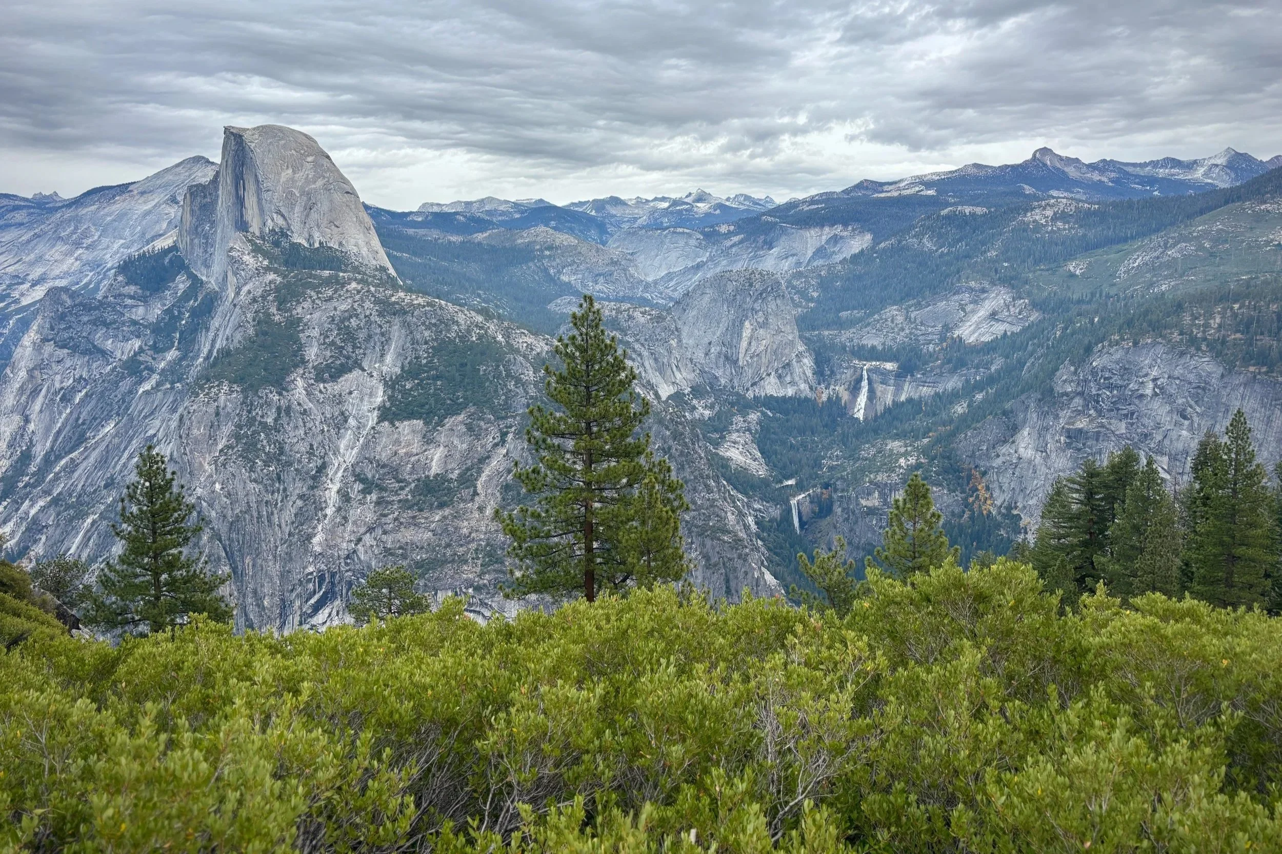 A scenic view of Yosemite National Park with granite cliffs, pine trees, and waterfalls under a cloudy sky.