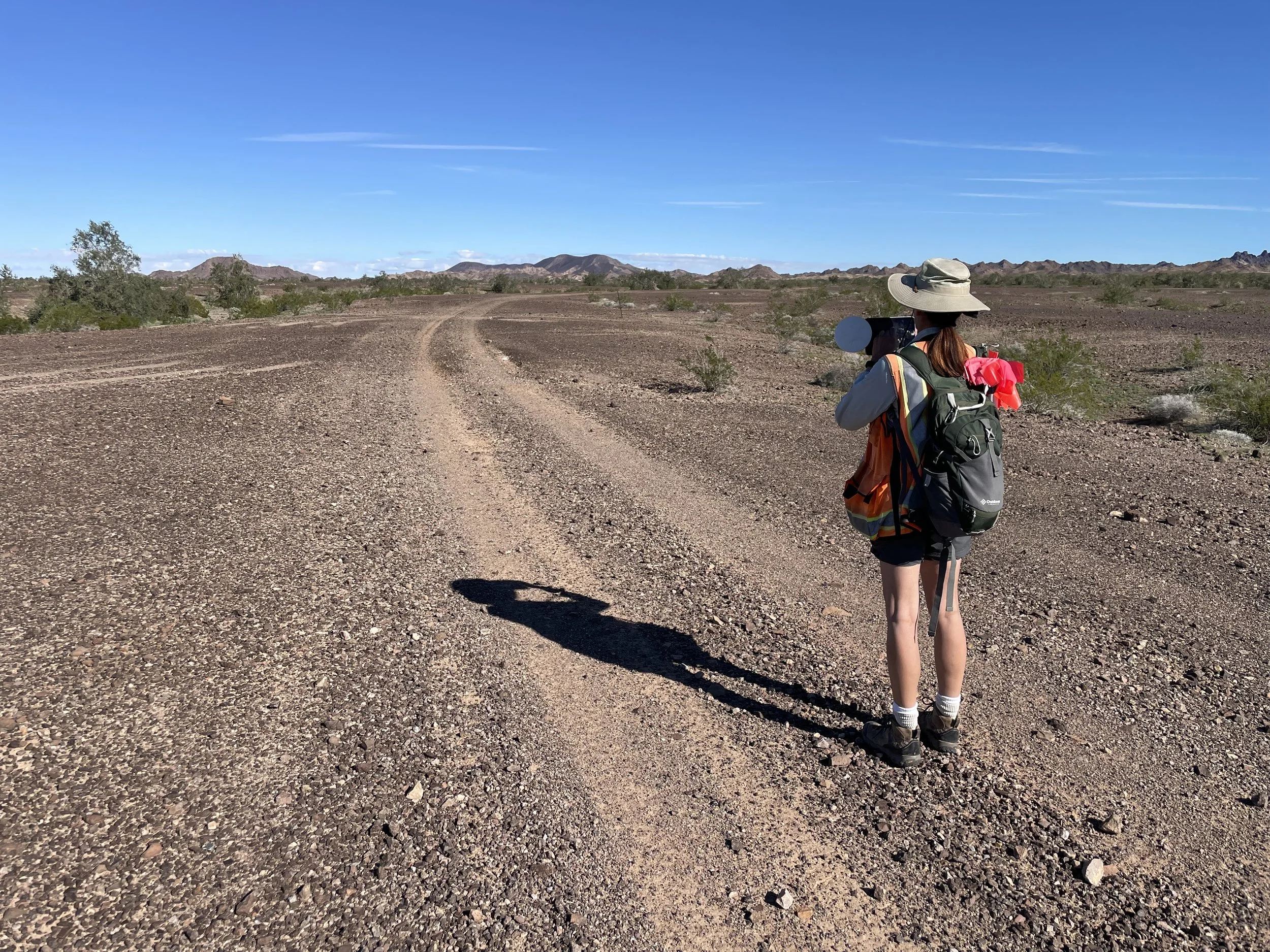 A woman with a hat, backpack, and hiking gear standing on a dirt road in an arid desert landscape.