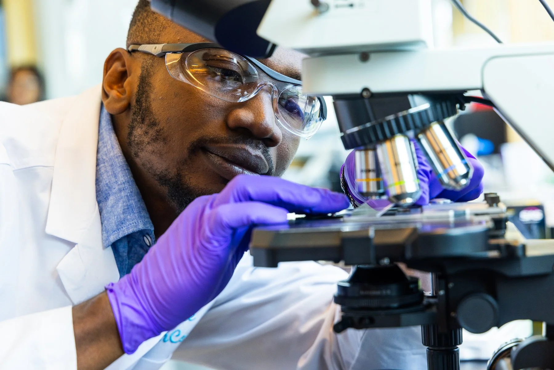 A scientist wearing safety glasses and purple gloves looking into a microscope in a laboratory.