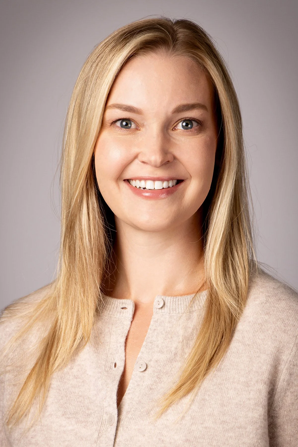 A woman with blonde hair smiling, wearing a beige sweater with buttons, against a plain gray background. Salt Lake City Portrait & Headshot Photographer - Nick Sokoloff - Utah