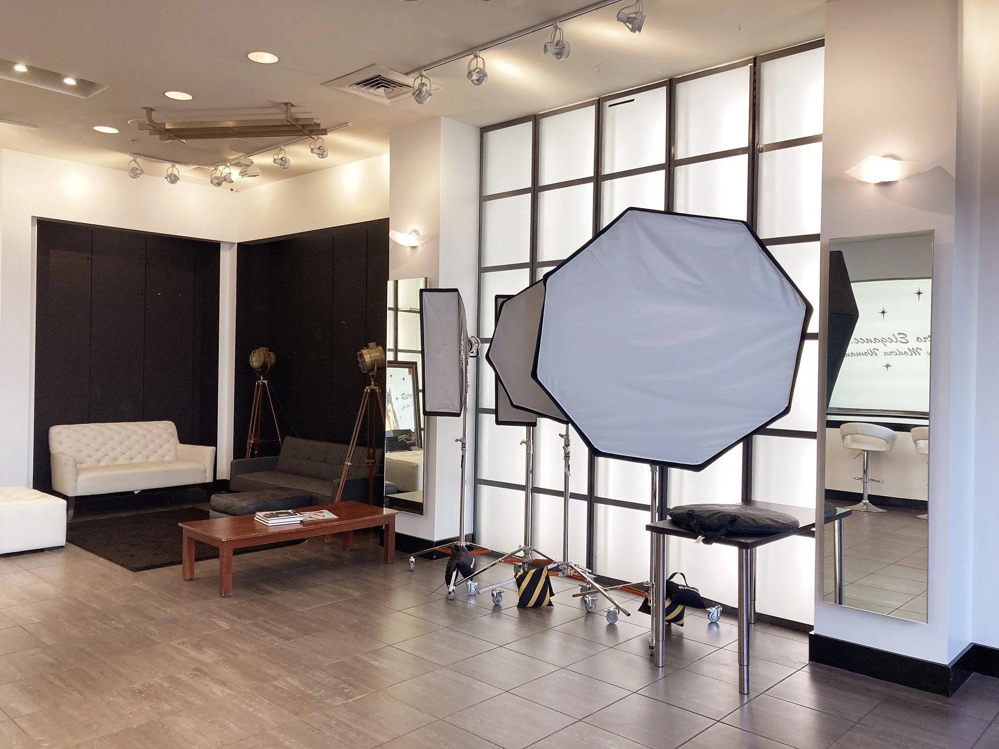 Indoor photo of a photography studio setup with lighting equipment and sofas. There are large softbox lights, a tripod with stage lights, a mirror, and a small seating area with a white and a gray sofa, a coffee table, and a black rug. The room has m