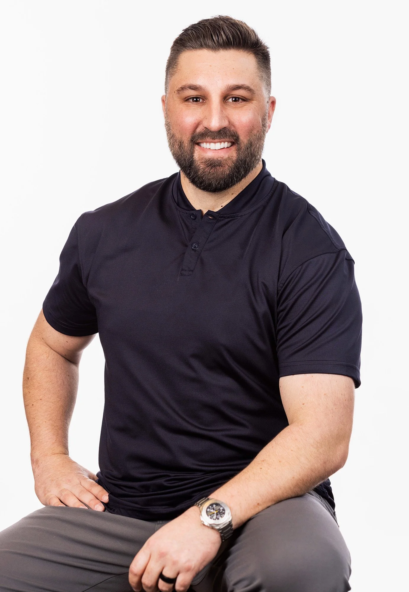 A smiling man with a beard, wearing a black short-sleeve polo shirt, seated against a white background.