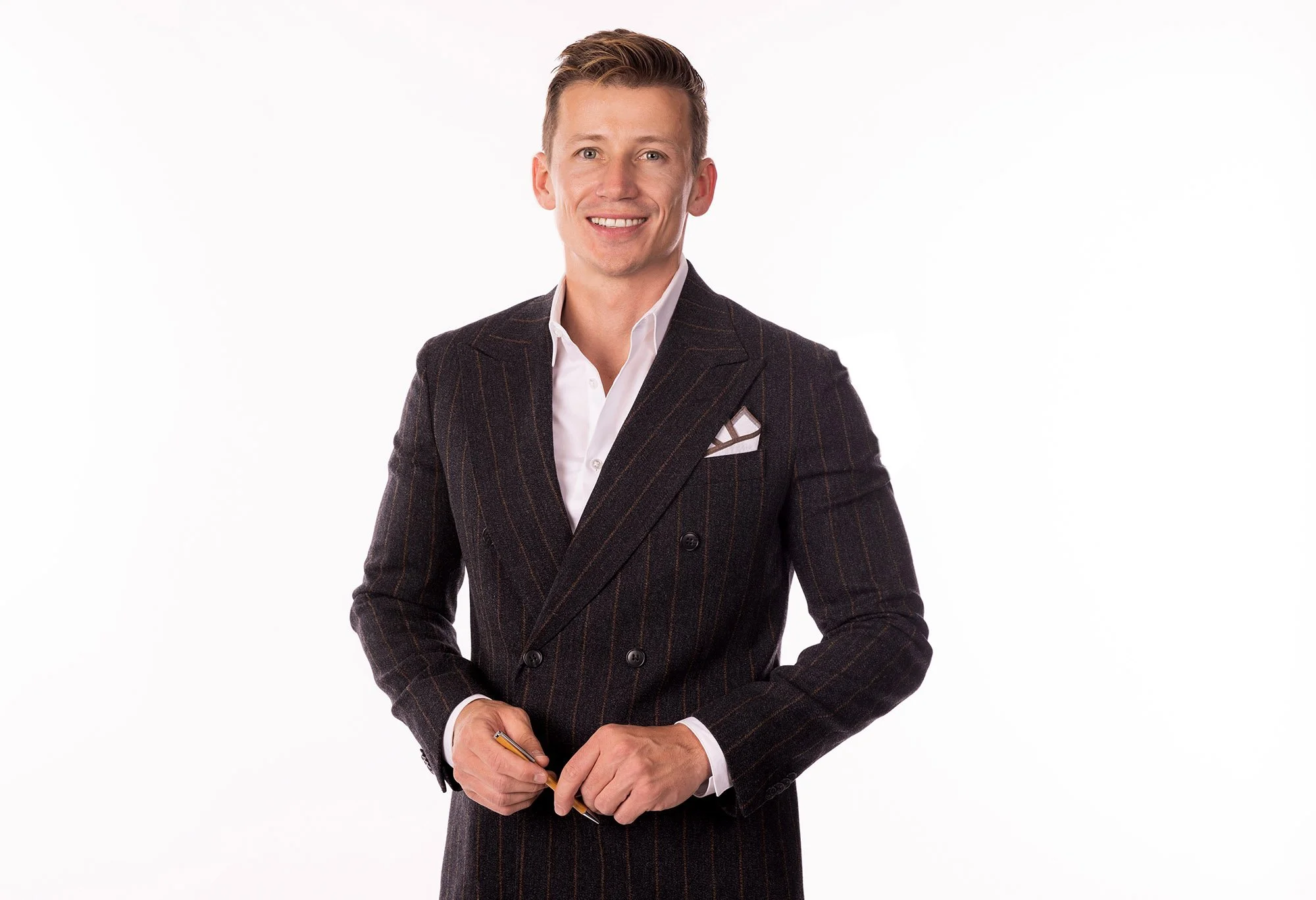 A young man with blonde hair, dressed in a dark, striped double-breasted suit, white shirt, and pocket square, smiling while holding a pen against a white background. Salt Lake City Portrait & Headshot Photographer - Nick Sokoloff - Utah
