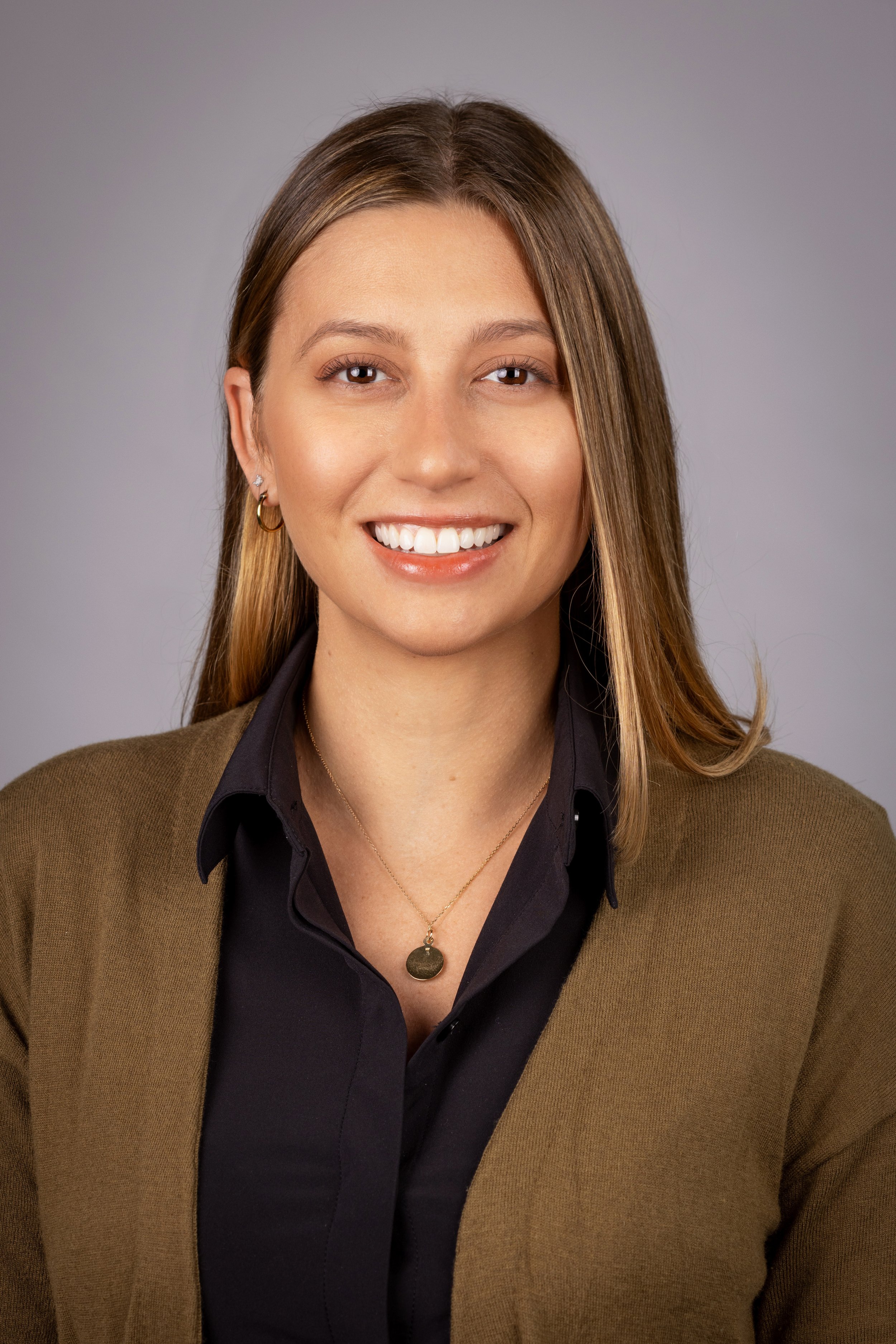 Salt Lake City Portrait & Headshot Photographer - Nick Sokoloff - Utah A smiling woman with long straight hair, wearing a black shirt, a brown blazer, and jewelry, against a plain gray background.