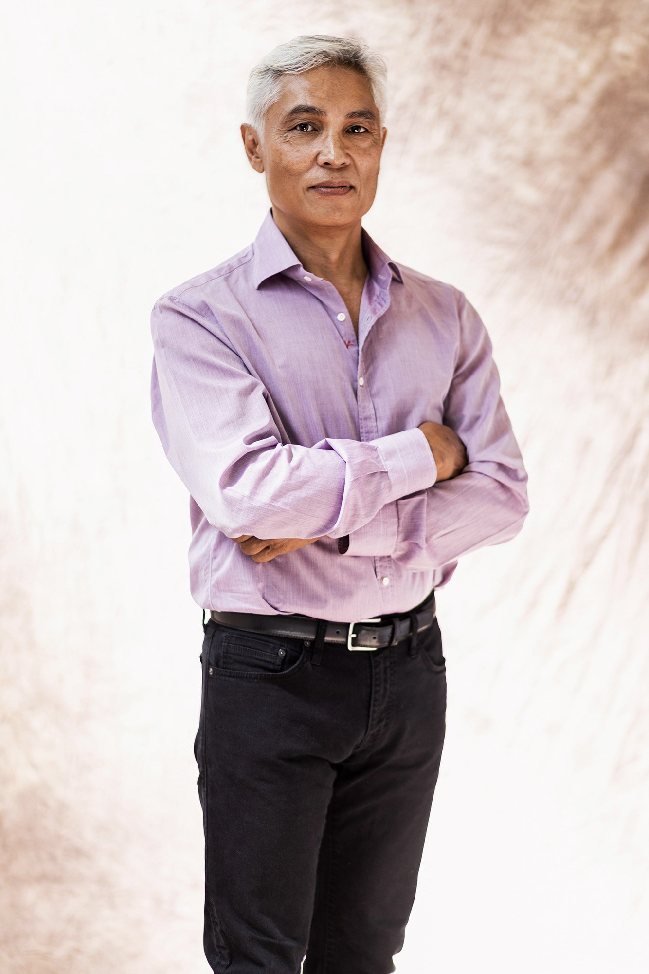 A man with gray hair wearing a lavender dress shirt and black jeans, standing with arms crossed against a neutral background.