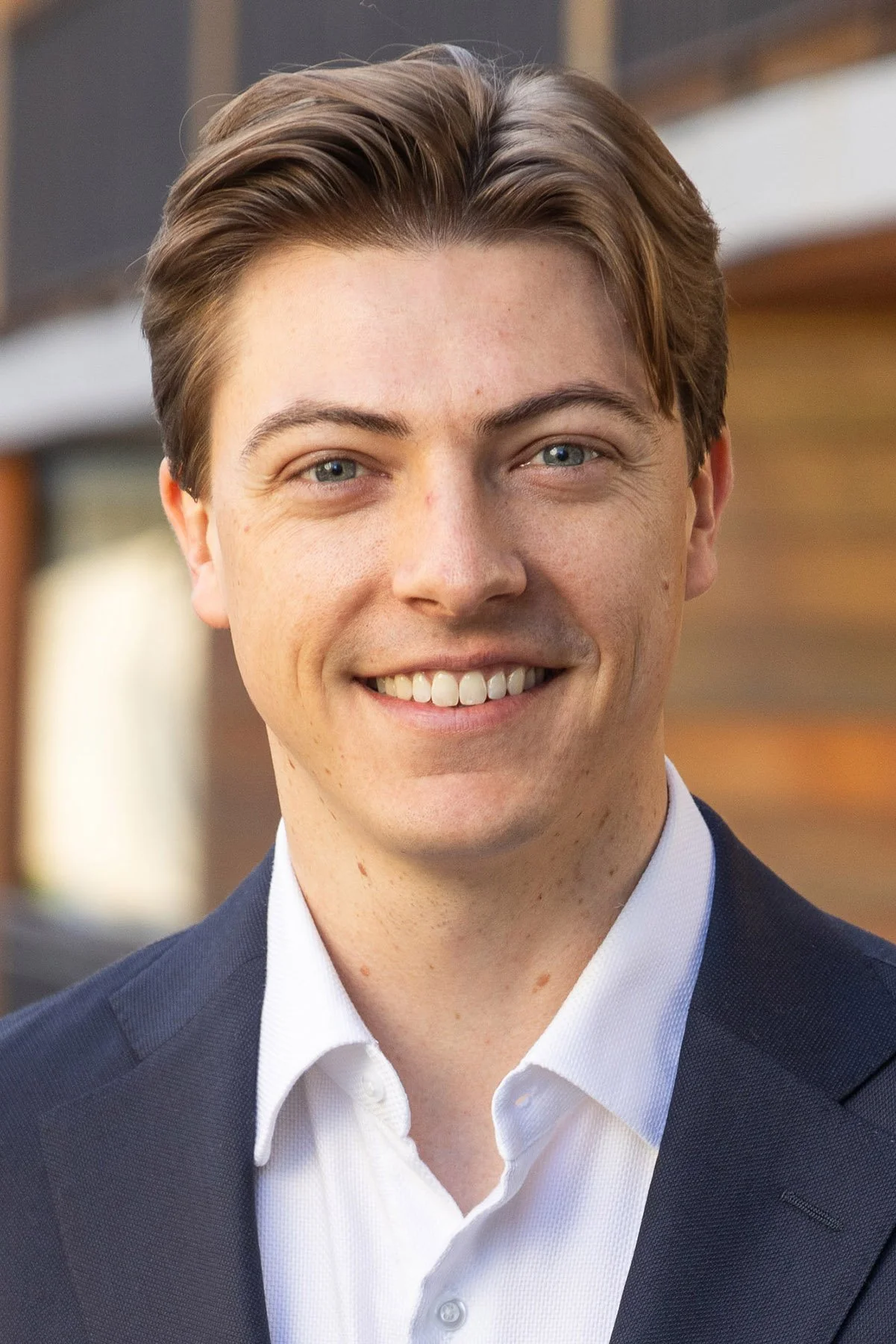 A young man with light skin, brown hair, blue eyes, and a friendly smile, wearing a navy suit and a white shirt, standing outdoors with a blurred background of a building.