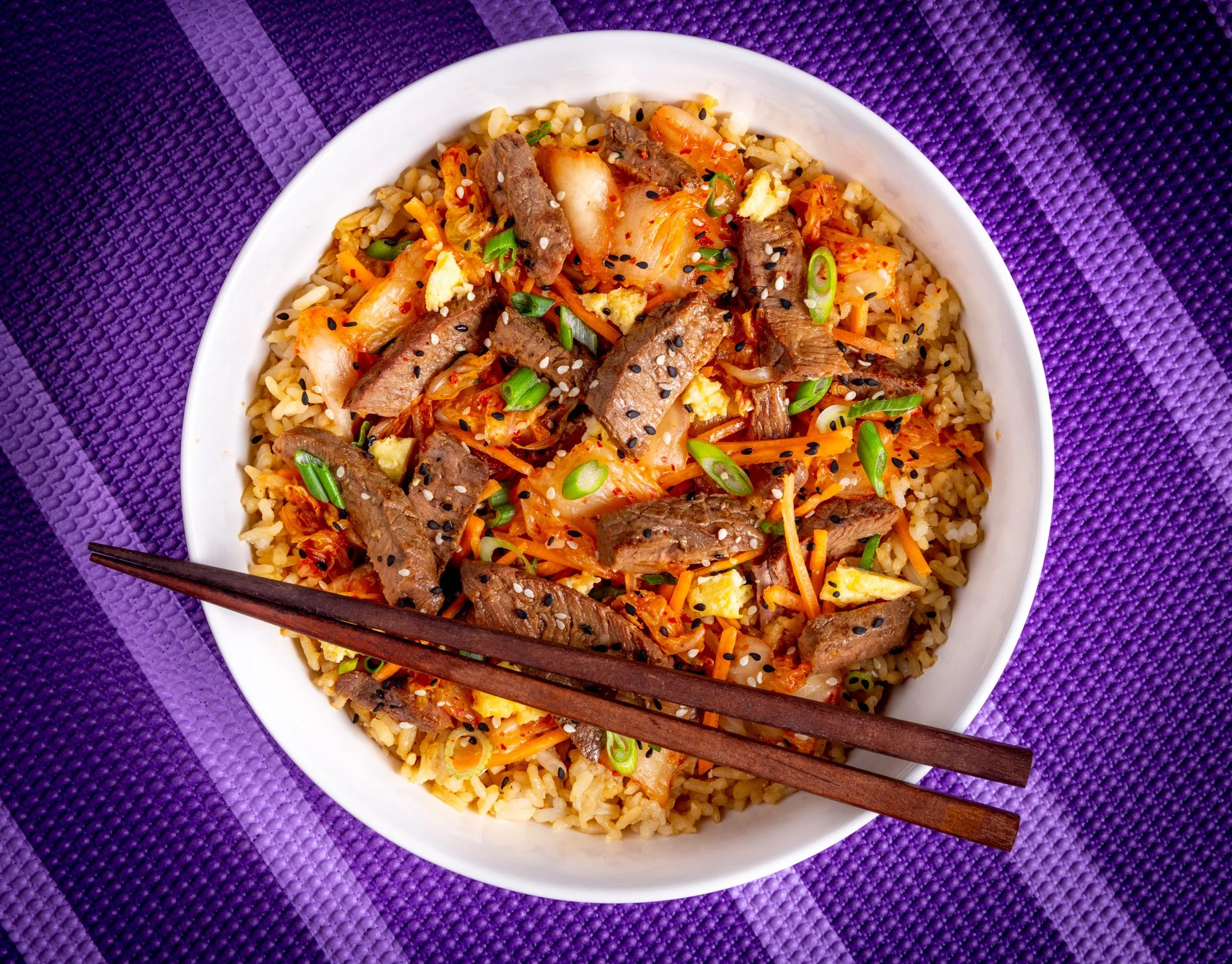 A white bowl of fried rice topped with beef, vegetables, and sesame seeds, placed on a purple textured surface with chopsticks across the bowl.