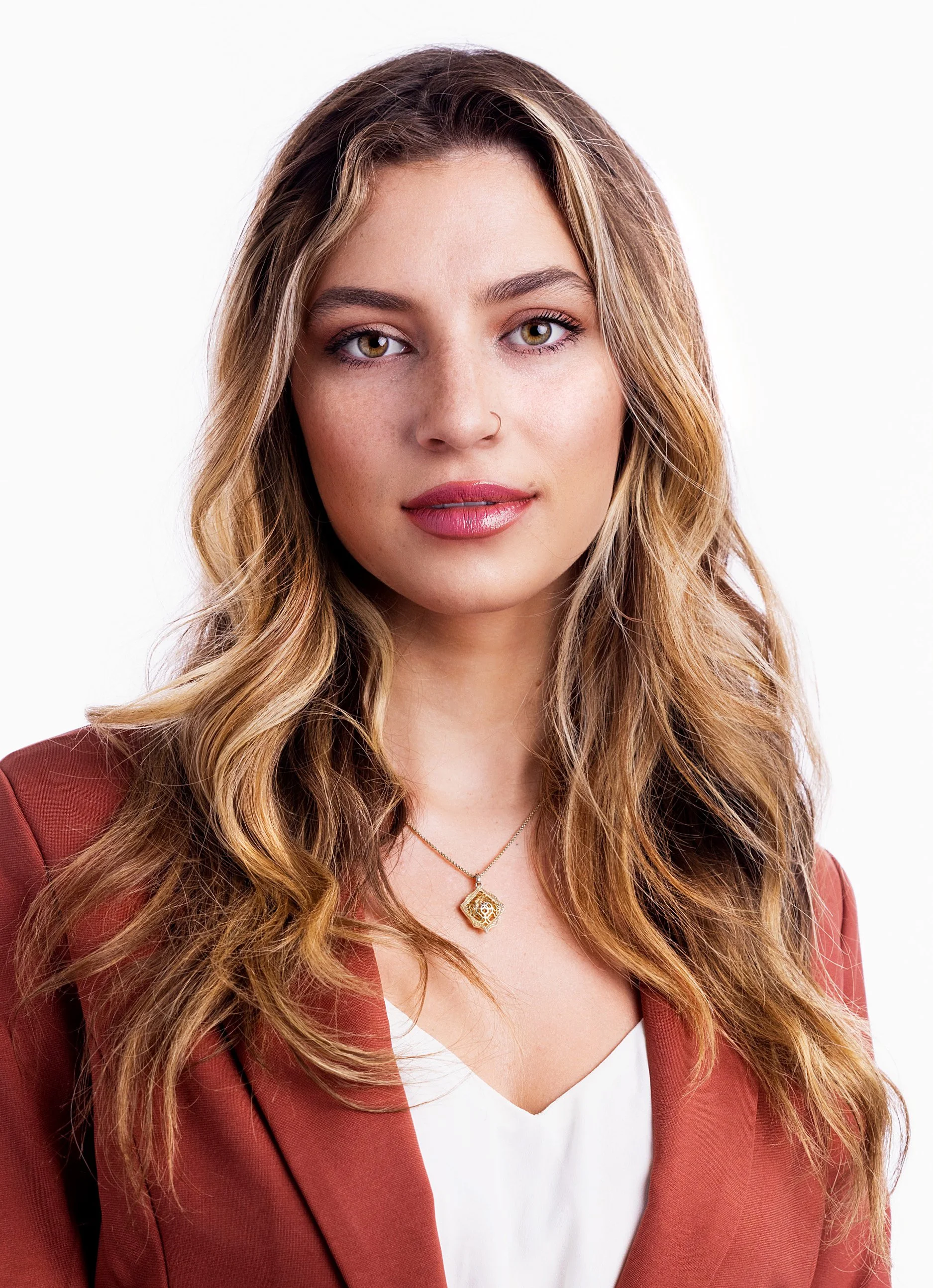 A young woman with long wavy light brown hair, hazel eyes, wearing a rust-colored blazer over a white top, gold necklace with a decorative pendant, and makeup with pink lipstick, posing against a plain white background.