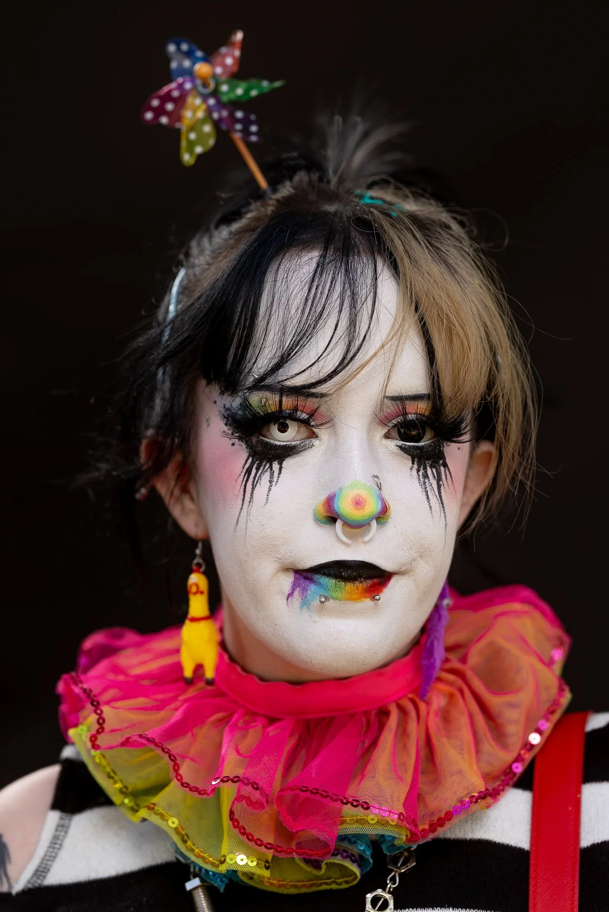 A person with clown makeup and colorful accessories, including a rainbow-colored ruffle collar, a rainbow-colored clown nose, and rainbow teaser earrings, against a black background.