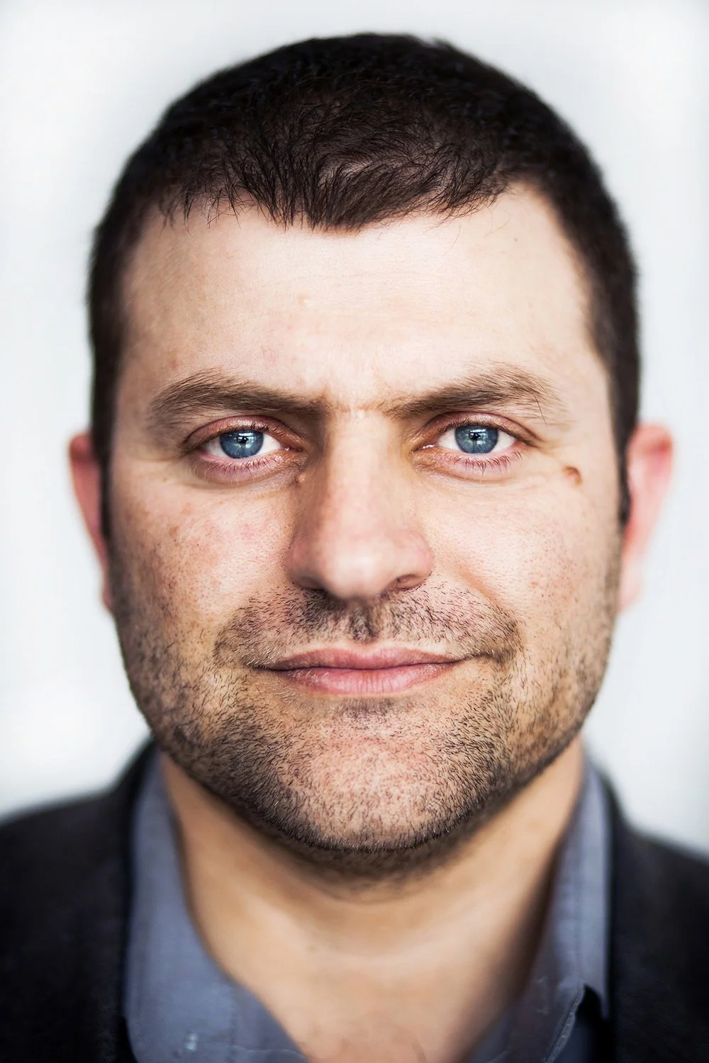 Close-up of a man with short dark hair, blue eyes, light stubble, and a slight smile, wearing a dark blazer and gray shirt.