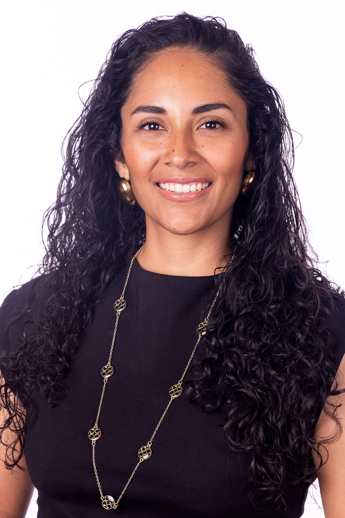 A woman with long, curly dark hair, wearing gold hoop earrings, a black top, and a gold necklace, smiling at the camera. Salt Lake City Portrait & Headshot Photographer - Nick Sokoloff - Utah