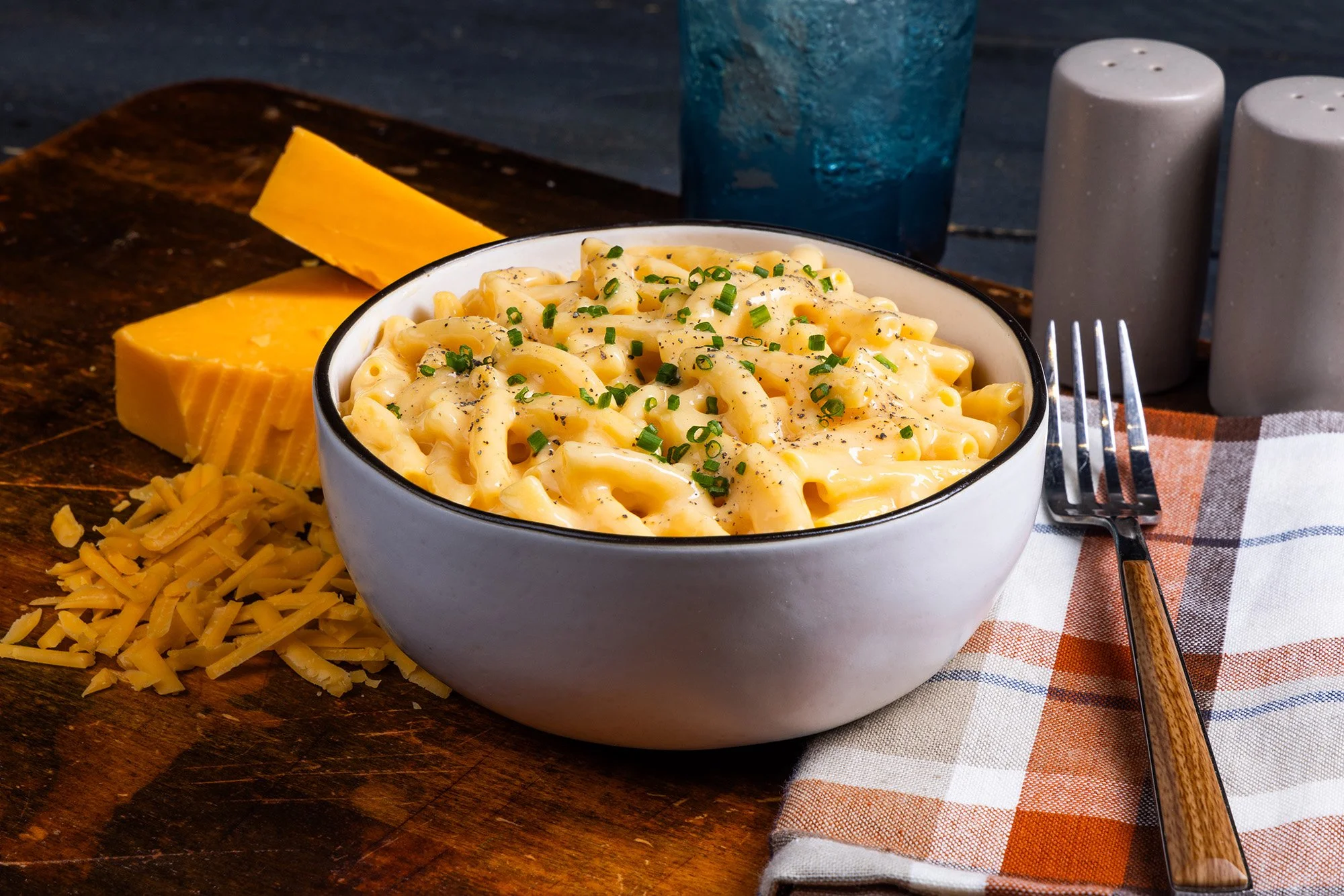 A bowl of creamy macaroni and cheese garnished with chopped green herbs on a wooden table, with shredded cheese, slices of cheese, a blue drink, salt and pepper shakers, and a fork on a plaid napkin.