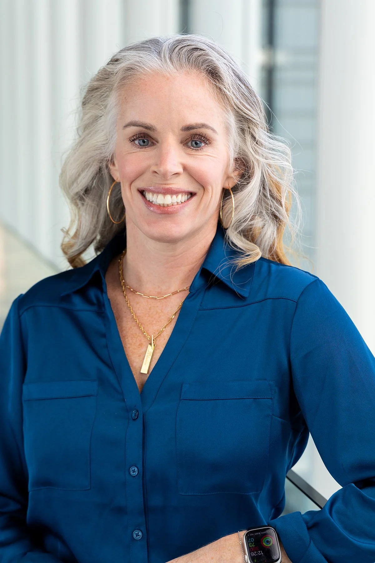 A smiling middle-aged woman with gray hair wearing a blue blouse, gold hoop earrings, layered necklaces, and an Apple Watch, standing in a bright indoor space with large windows in the background. Salt Lake City Portrait & Headshot Photographer - Nic