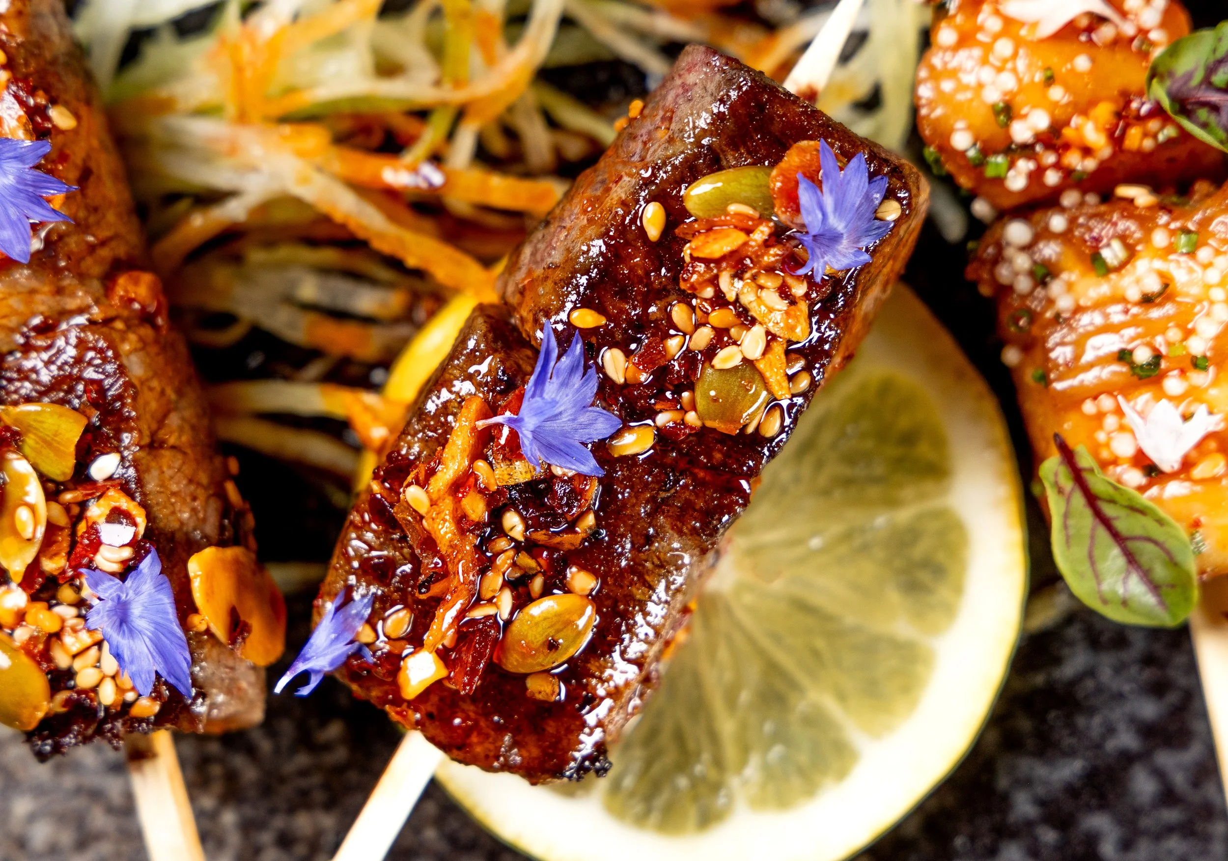 Close-up of grilled skewers with glazed meat, topped with seeds, spices, and edible blue flowers, garnished with lemon slices and shredded vegetables.