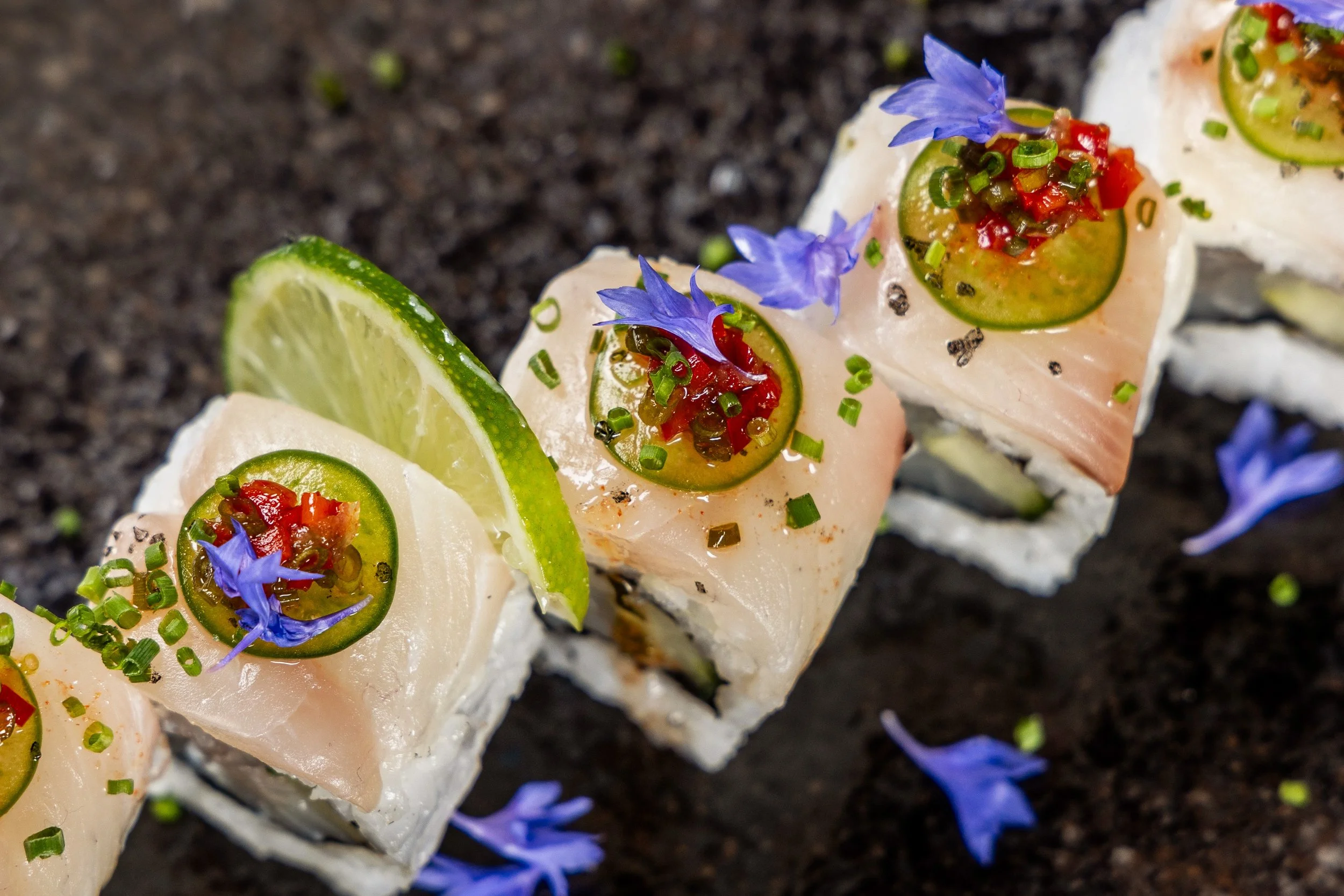 Close-up of sushi rolls topped with sliced fish, lime slices, jalapeño slices, chopped green onions, red chili bits, and blue edible flowers on a dark surface