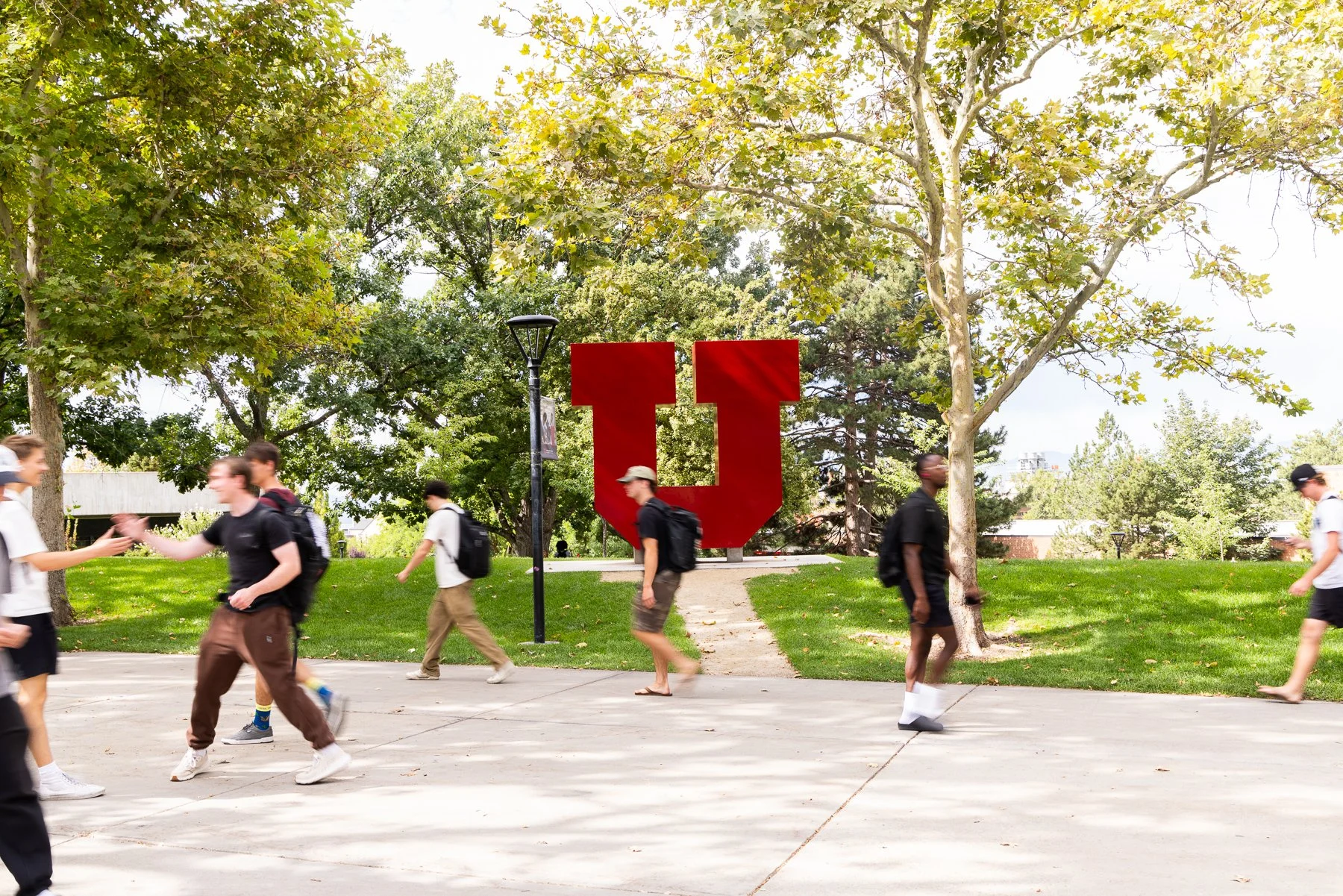 Students walking and greeting each other on a college campus with a large red university sign in the background, surrounded by trees and greenery.