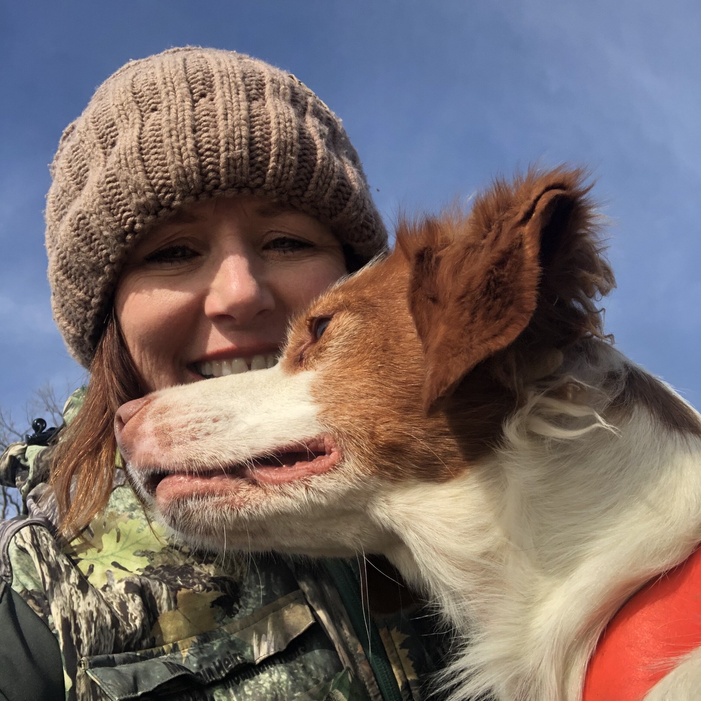 Person wearing a knit hat and camouflage jacket, smiling with a brown and white dog outdoors.