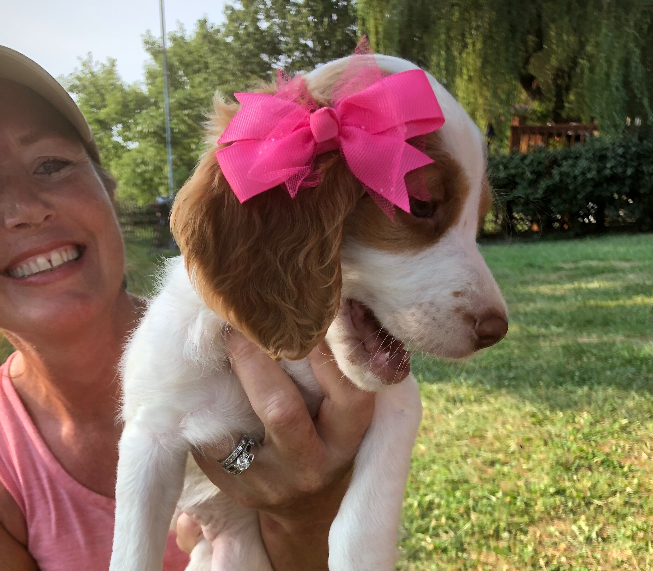 A woman holding a puppy wearing a pink bow, with grassy background.