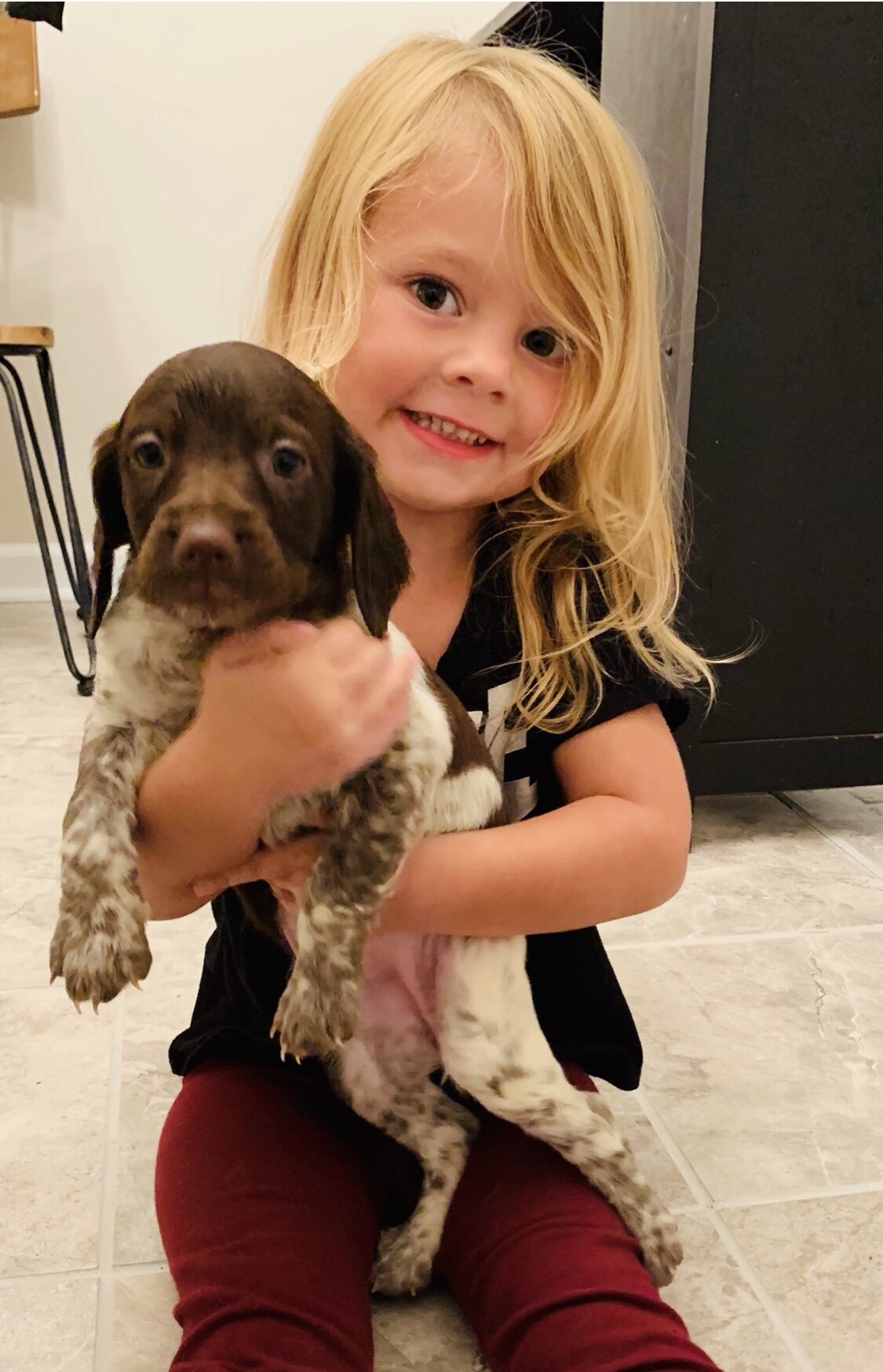 Smiling child holding a brown and white puppy indoors on a tiled floor.