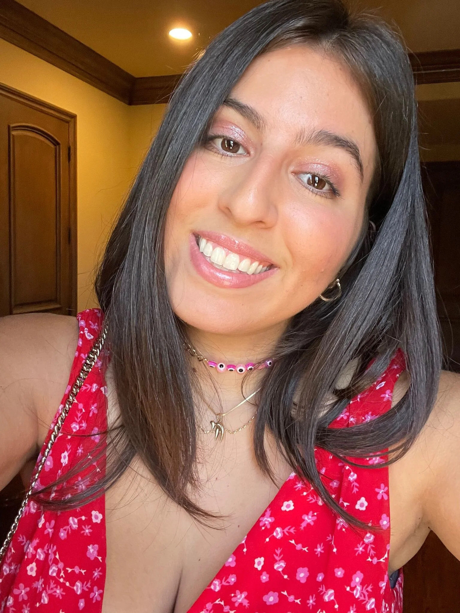 Close-up of a young woman with long dark hair, smiling, wearing a red floral dress, layered necklaces, and a pink beaded choker.