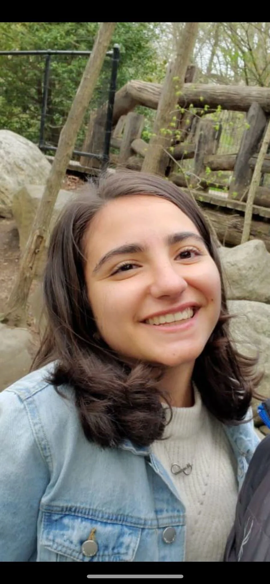 A young woman smiling outdoors, surrounded by trees and rocks, with a wooden and metal railing in the background.