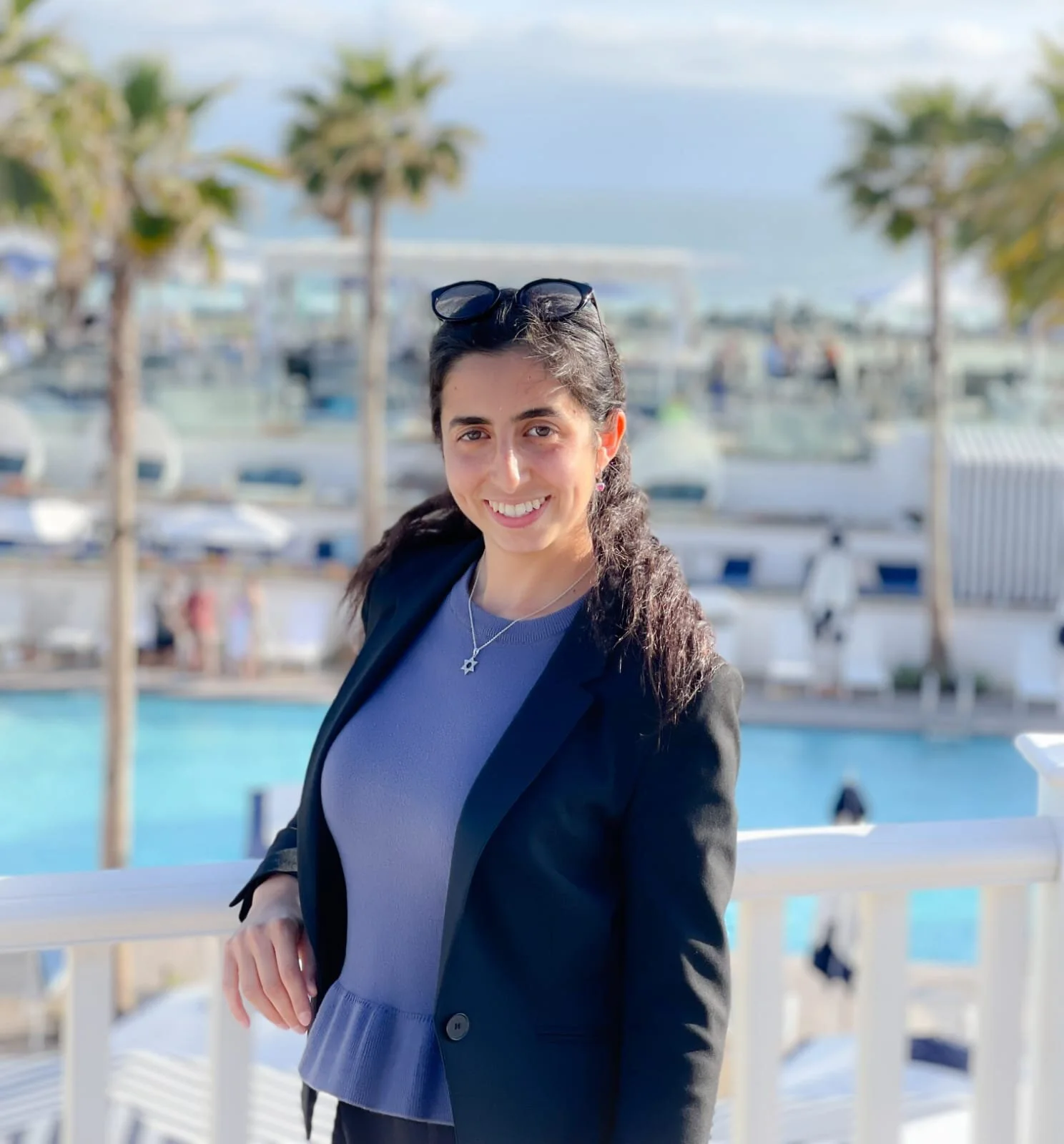 A smiling woman in a black blazer and purple top standing outdoors near a pool, with palm trees and a marina in the background.