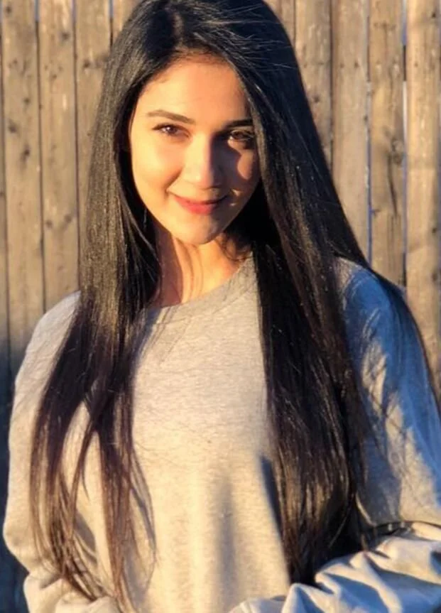 A young woman with long black hair smiling outdoors in front of a wooden fence, wearing a beige sweater.