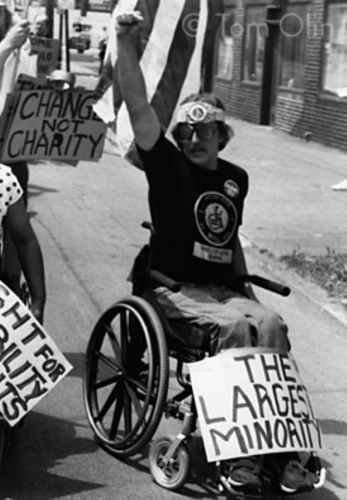 Black and white photo of person in a manual wheelchair holding up their fist. A sign resting on their legs reads "The Largest Minority," a posting in the back reads "Change not Charity" Photo taken by Tom Olin