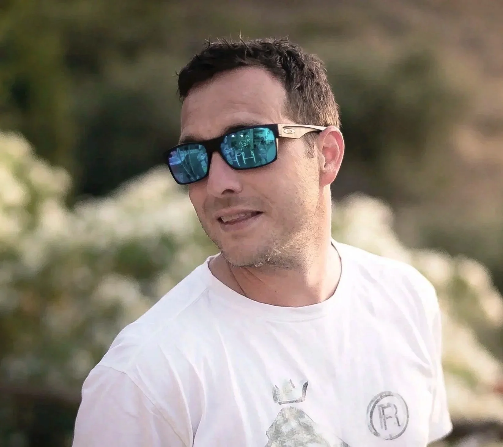 A man wearing reflective sunglasses and a white T-shirt outdoors with blurred trees and plants in the background.