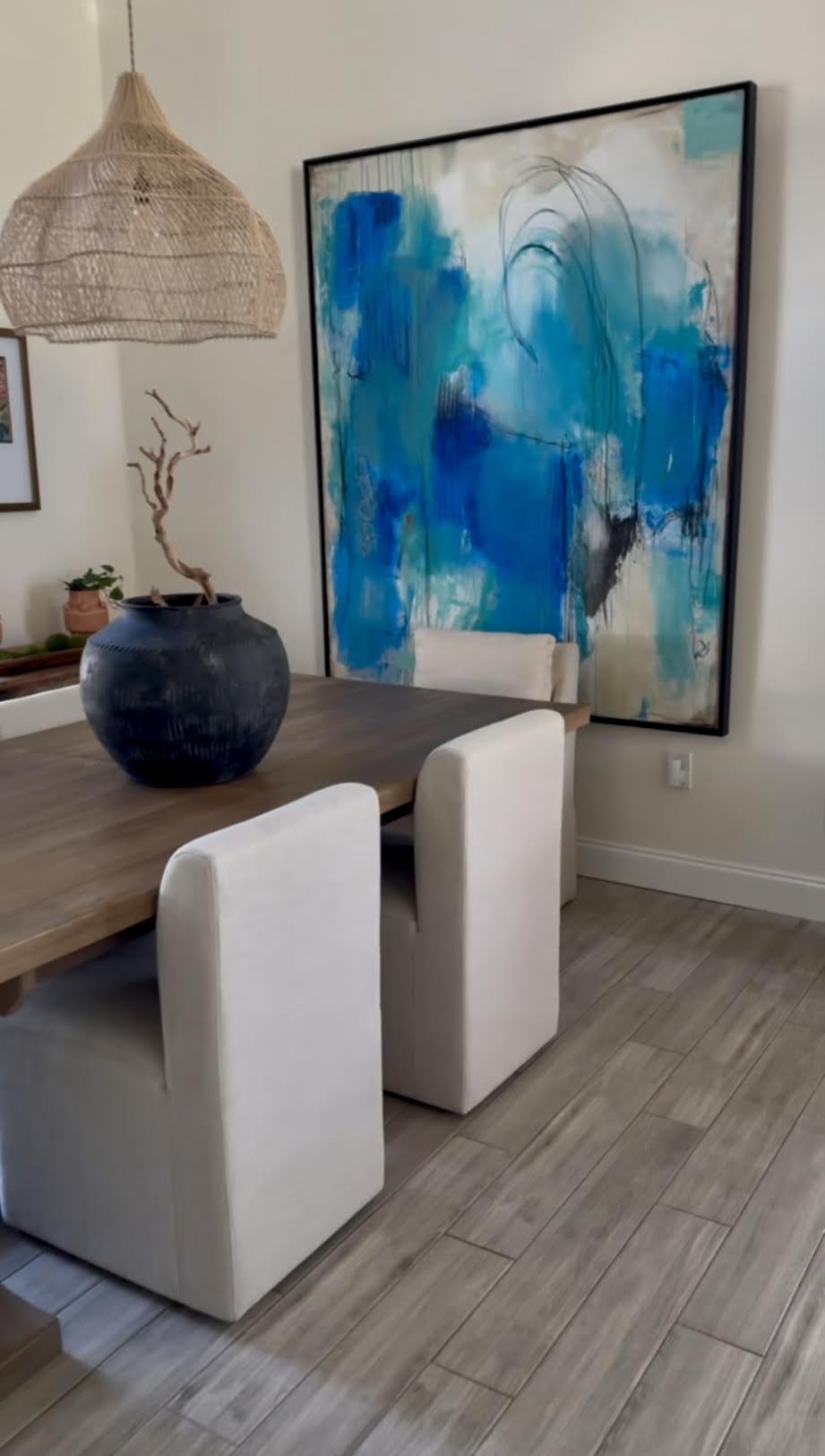 A dining room corner with a large, abstract blue painting, a wooden dining table with white upholstered chairs, a black pot with a dried tree branch, a woven pendant lamp, and a small potted plant.