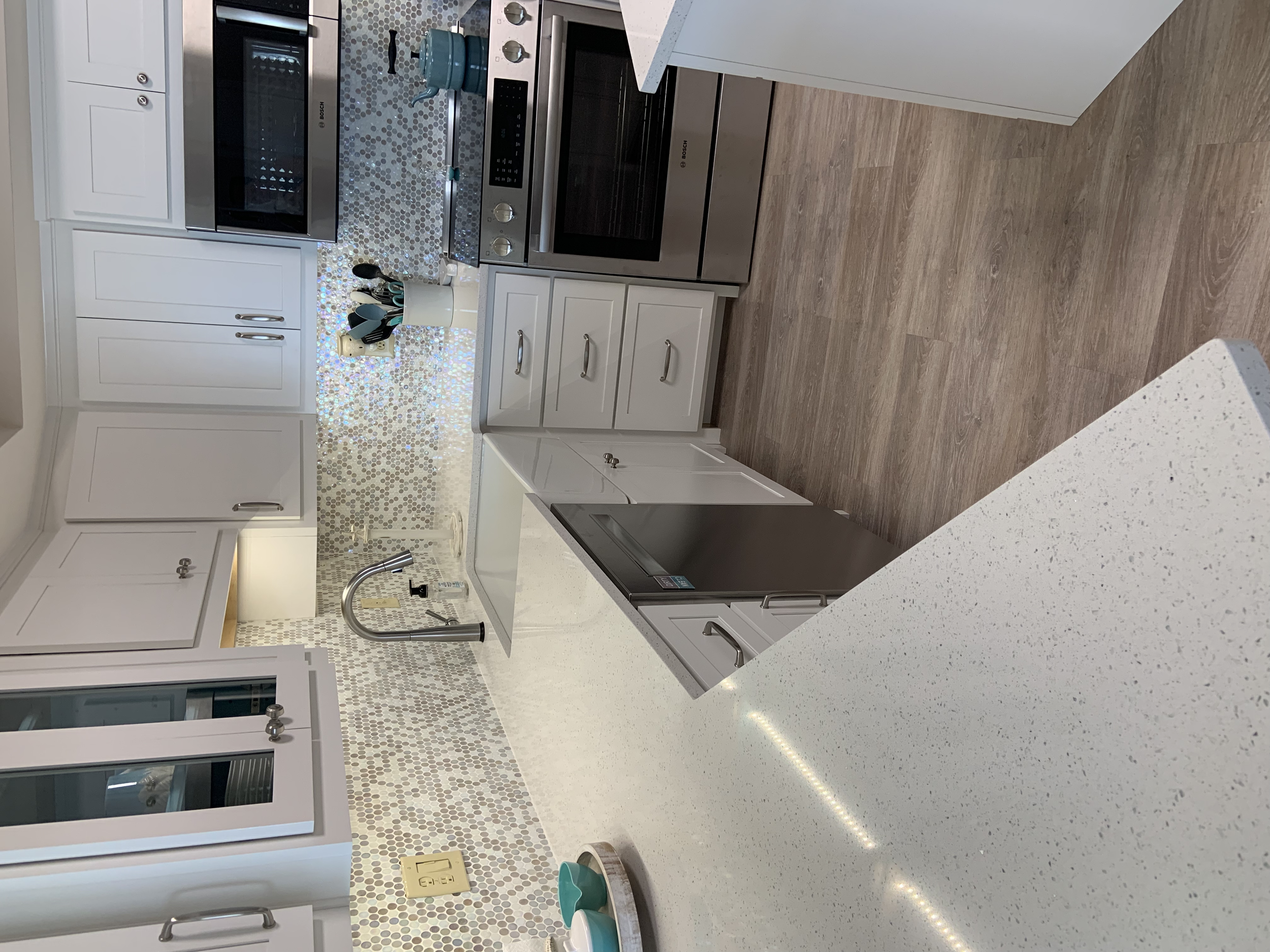A modern kitchen with white cabinetry, stainless steel appliances including an oven, microwave, and refrigerator. The backsplash features a mosaic tile pattern, and there is a kitchen island with a white countertop. A cup holding kitchen utensils is 