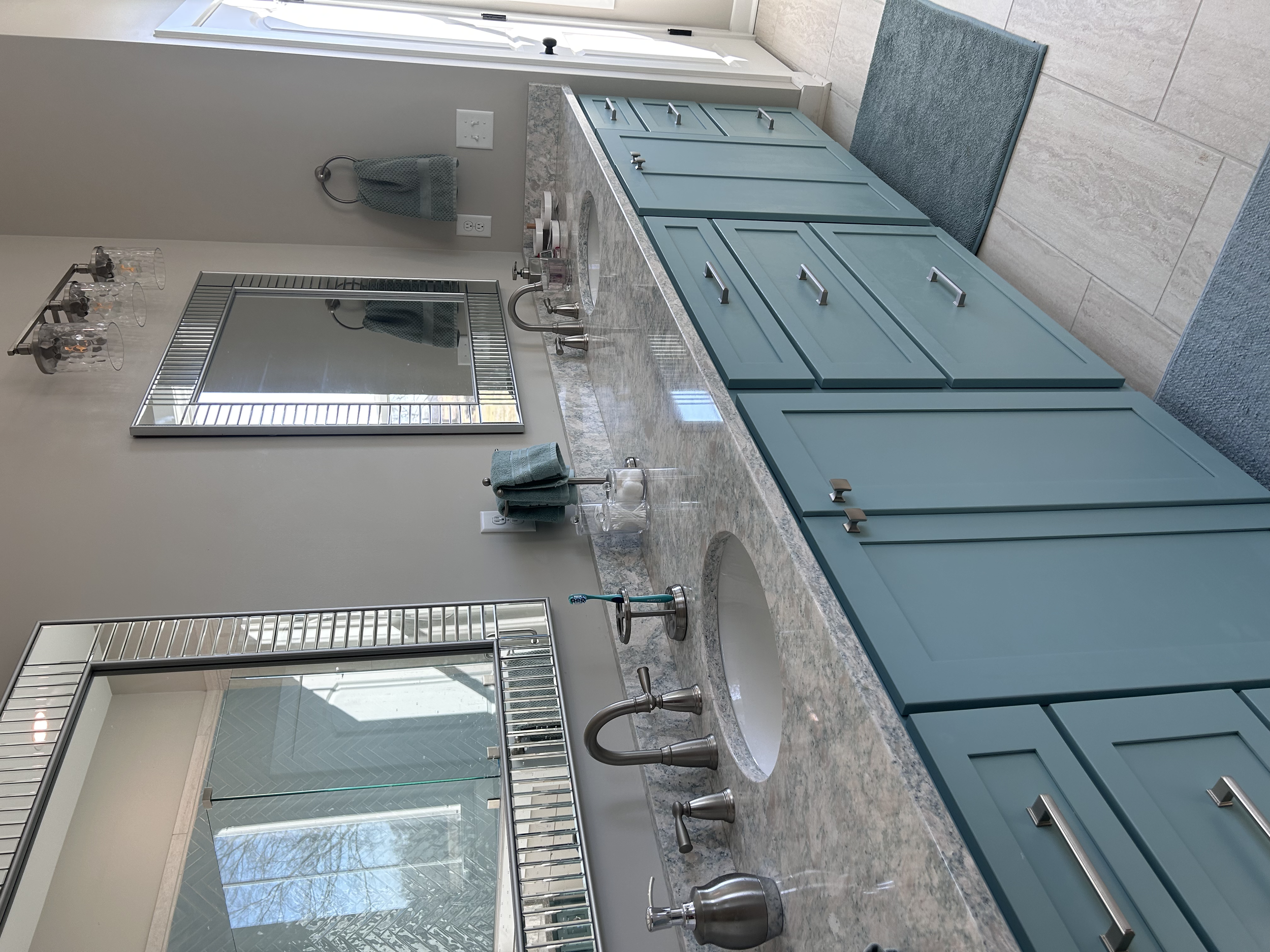 A bathroom vanity teal blue cabinets, a marble countertop, an oval sink, a mirror, and a window with a glass shower door nearby.
