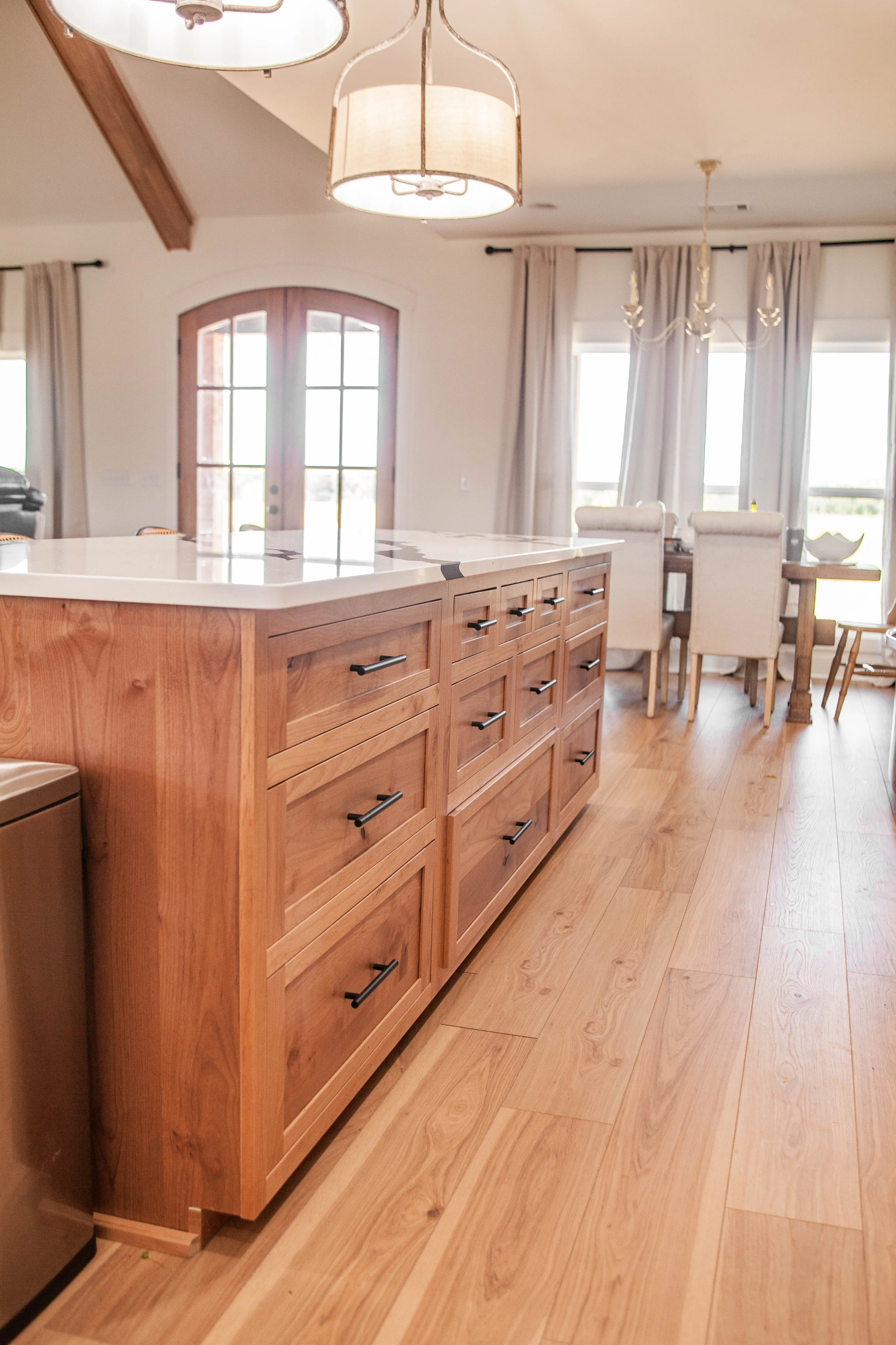 A spacious, well-lit kitchen and dining area with wooden flooring, a large island with multiple drawers, and a dining table with six chairs. The room features large windows with curtains, a door with glass panes, and two light fixtures hanging from t