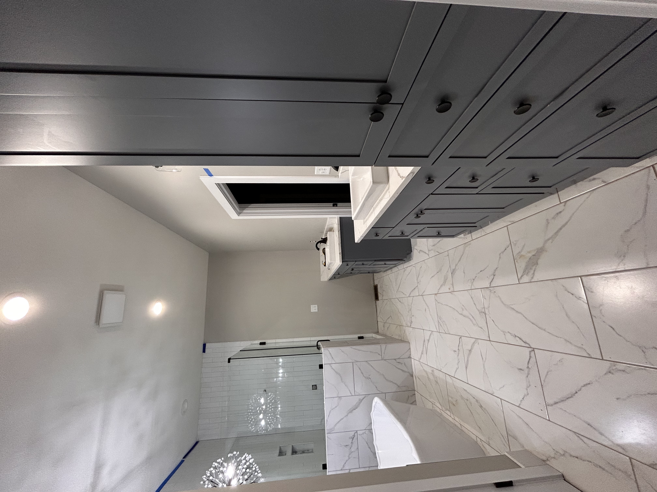 Modern bathroom with dark gray cabinetry, white marble countertops, and a marble floor. There is a window above the sink, and a glass shower in the background.