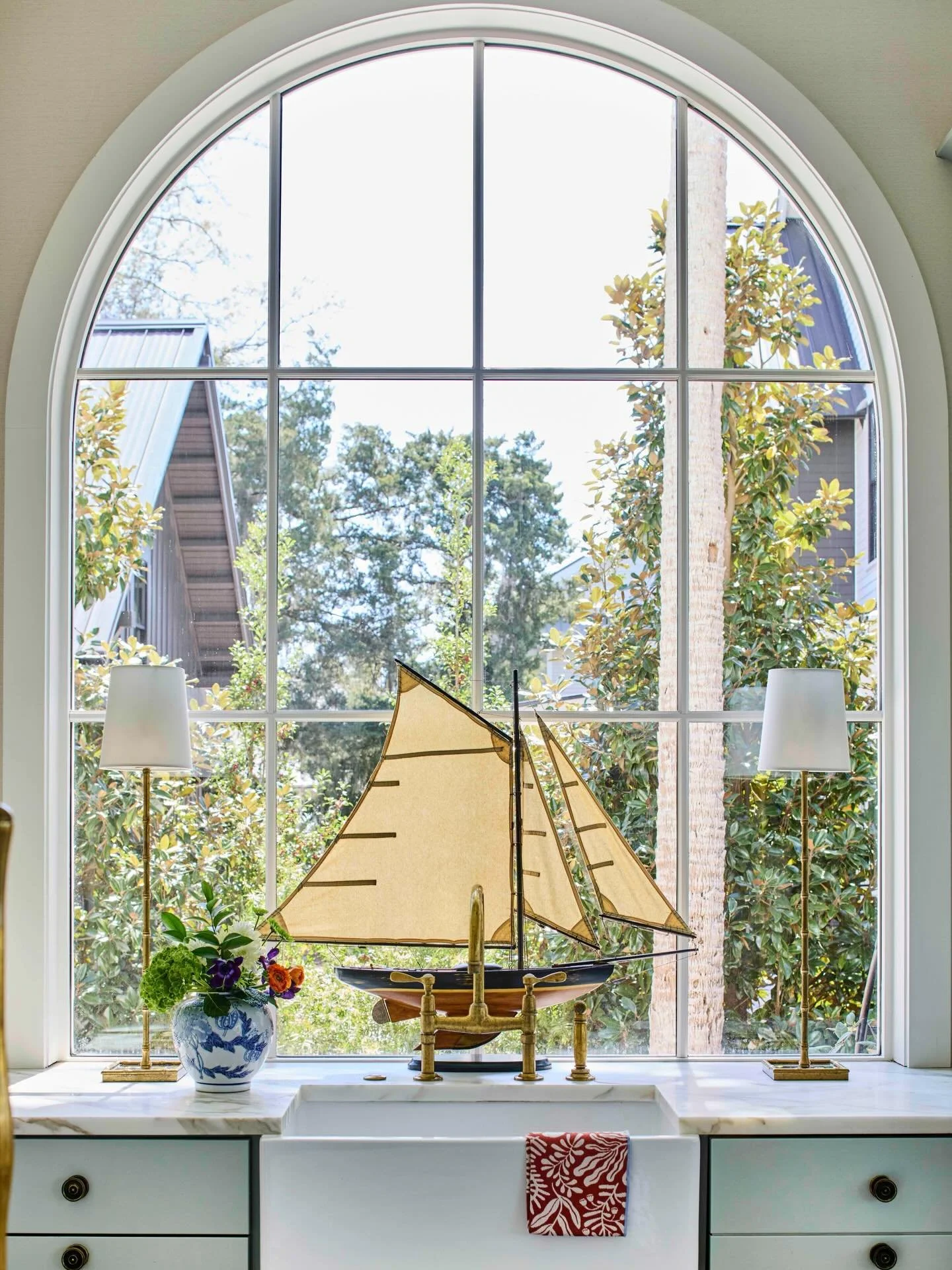 There&rsquo;s something magical about a window like this&mdash;sunlight pouring in, a view that feels like art, and just the right moment of whimsy sitting at the sink. A classic arched window, polished brass, and a sailing vessel ready to drift off 