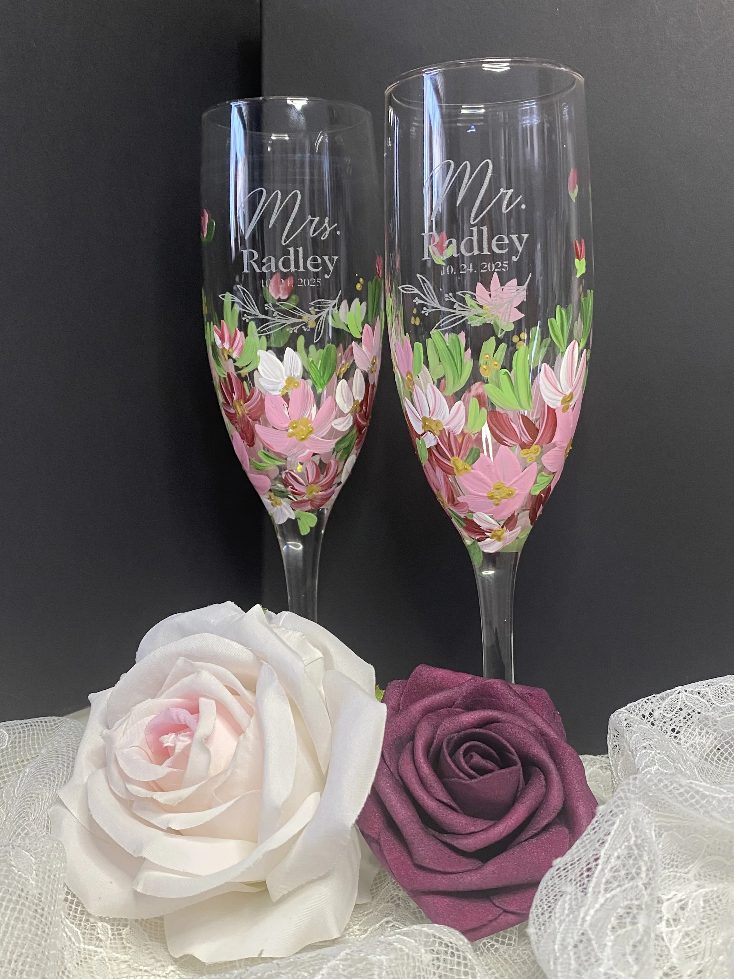 Two decorated champagne flutes with pink and green floral designs, labeled "Mrs. Radley" and "Mr. Radley" with the date October 24, 2025, placed on a lace fabric with a white and a purple artificial rose.