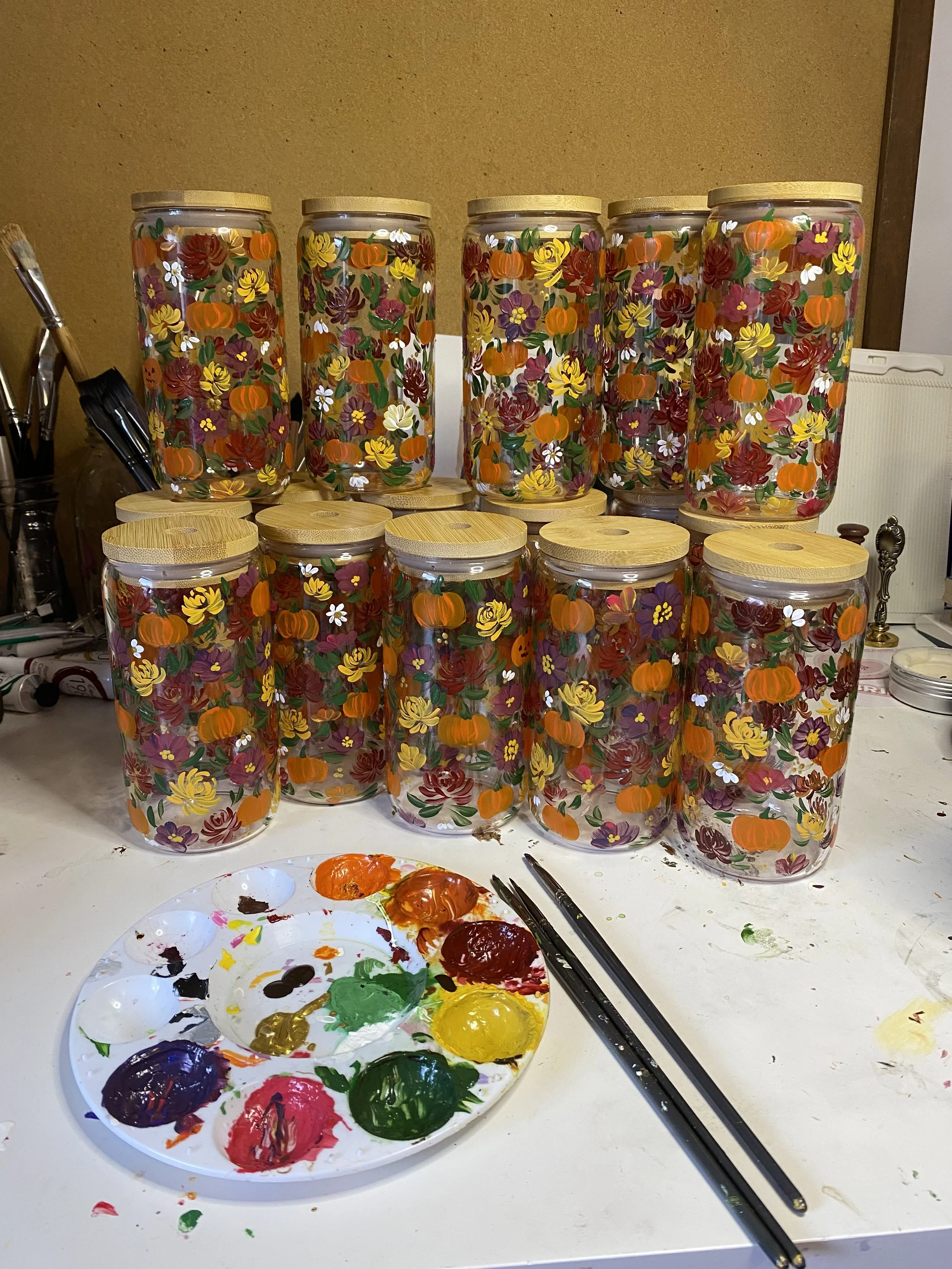 Ten painted glass jars with floral and pumpkin designs, wooden lids, on a work table with paint palette and brushes.