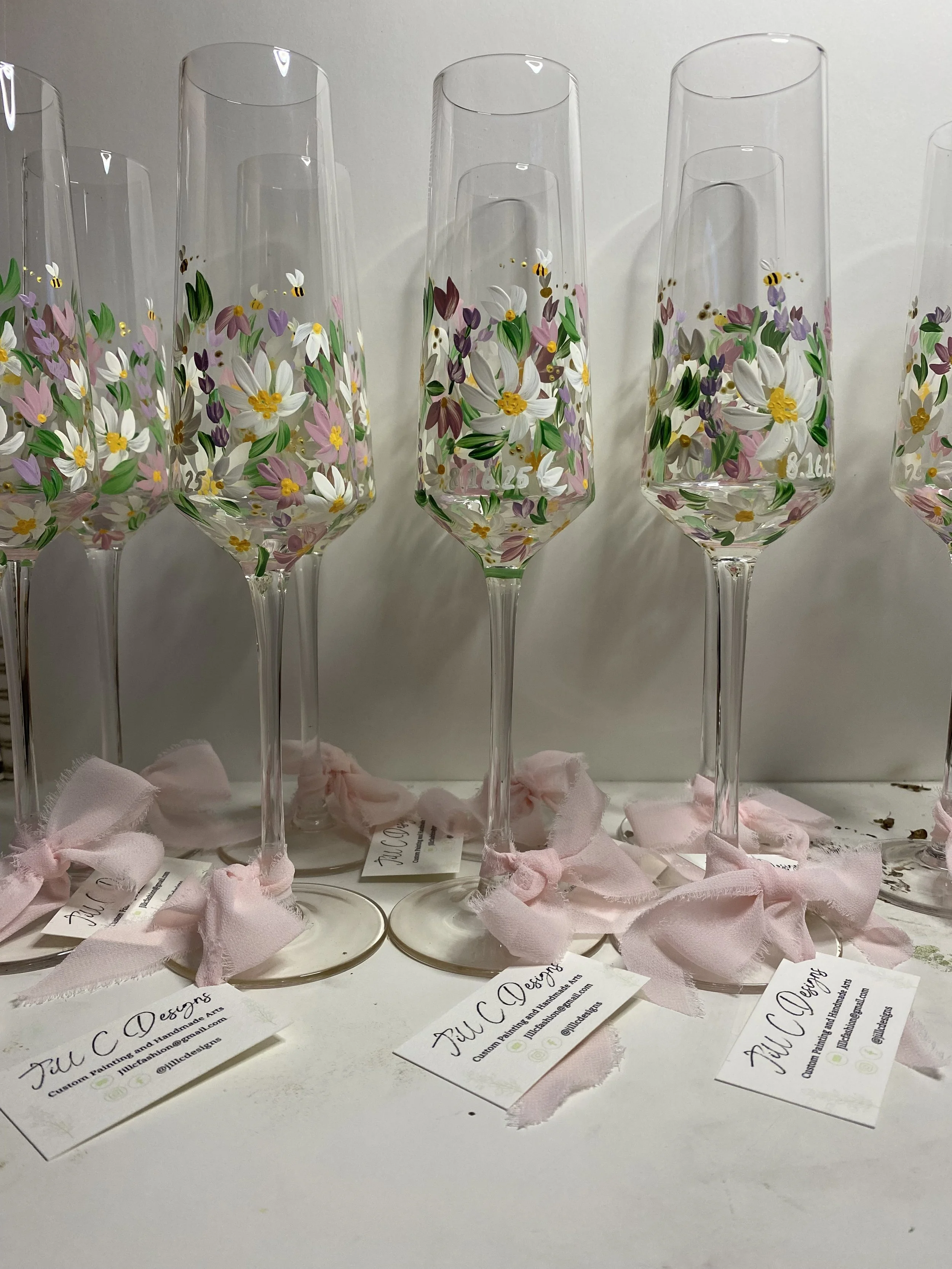 Set of tall champagne flutes decorated with floral designs and pink ribbons tied around the bases, with business cards reading "Jill C. Designs" placed in front.