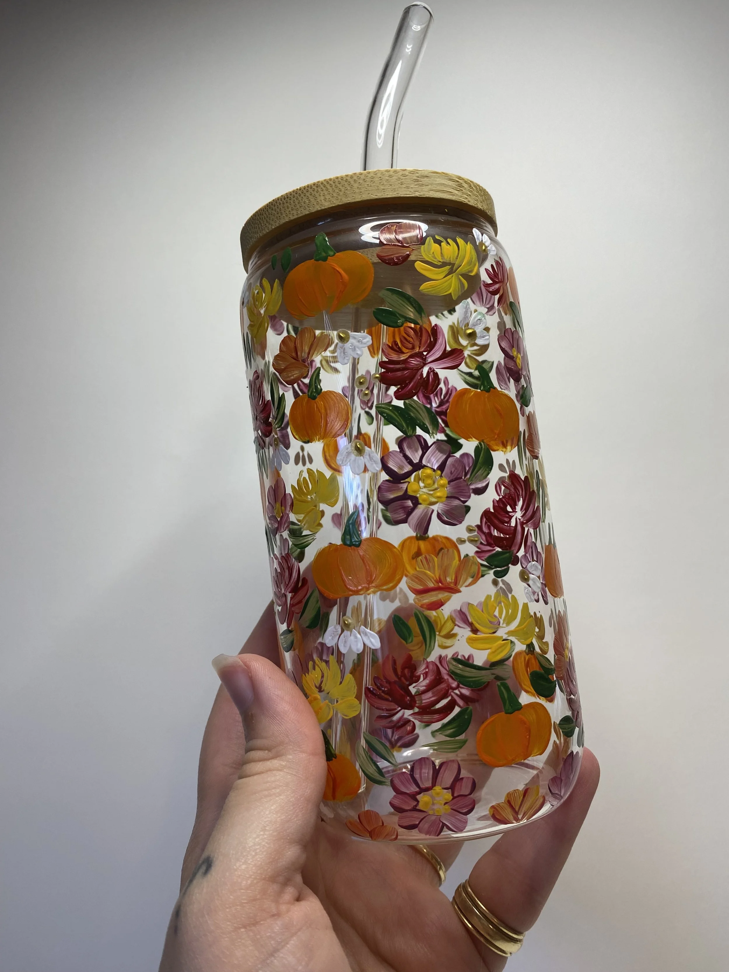 Hand holding a clear glass tumbler decorated with colorful painted flowers and pumpkins, with a bamboo lid and a straw.