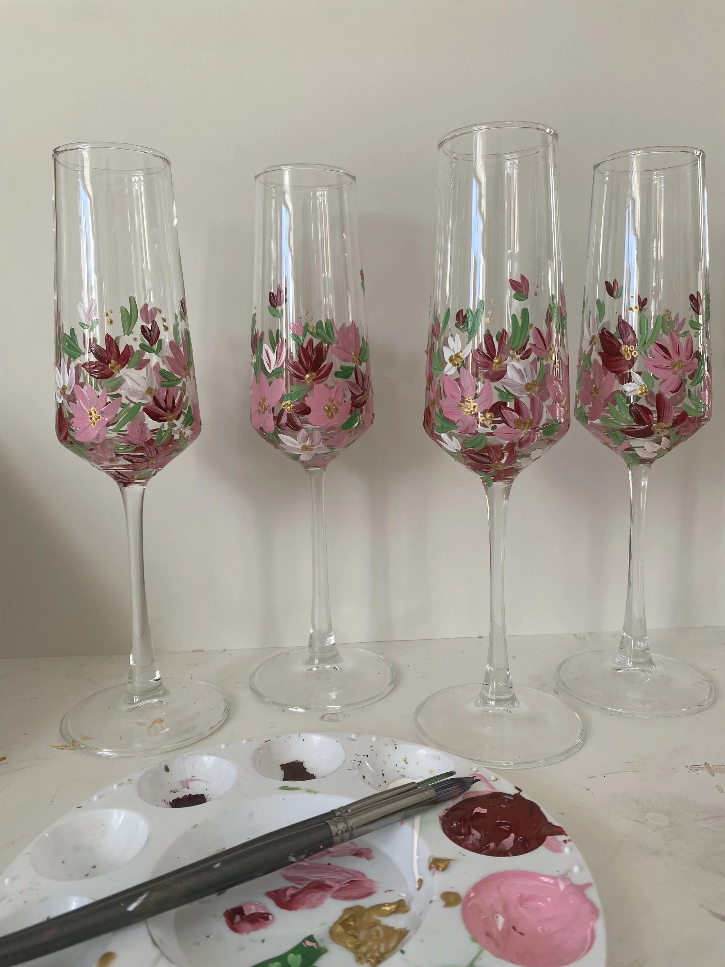 Four champagne flutes being hand-painted with pink, red, green, white, and gold floral designs, with a paint palette and brushes in front.