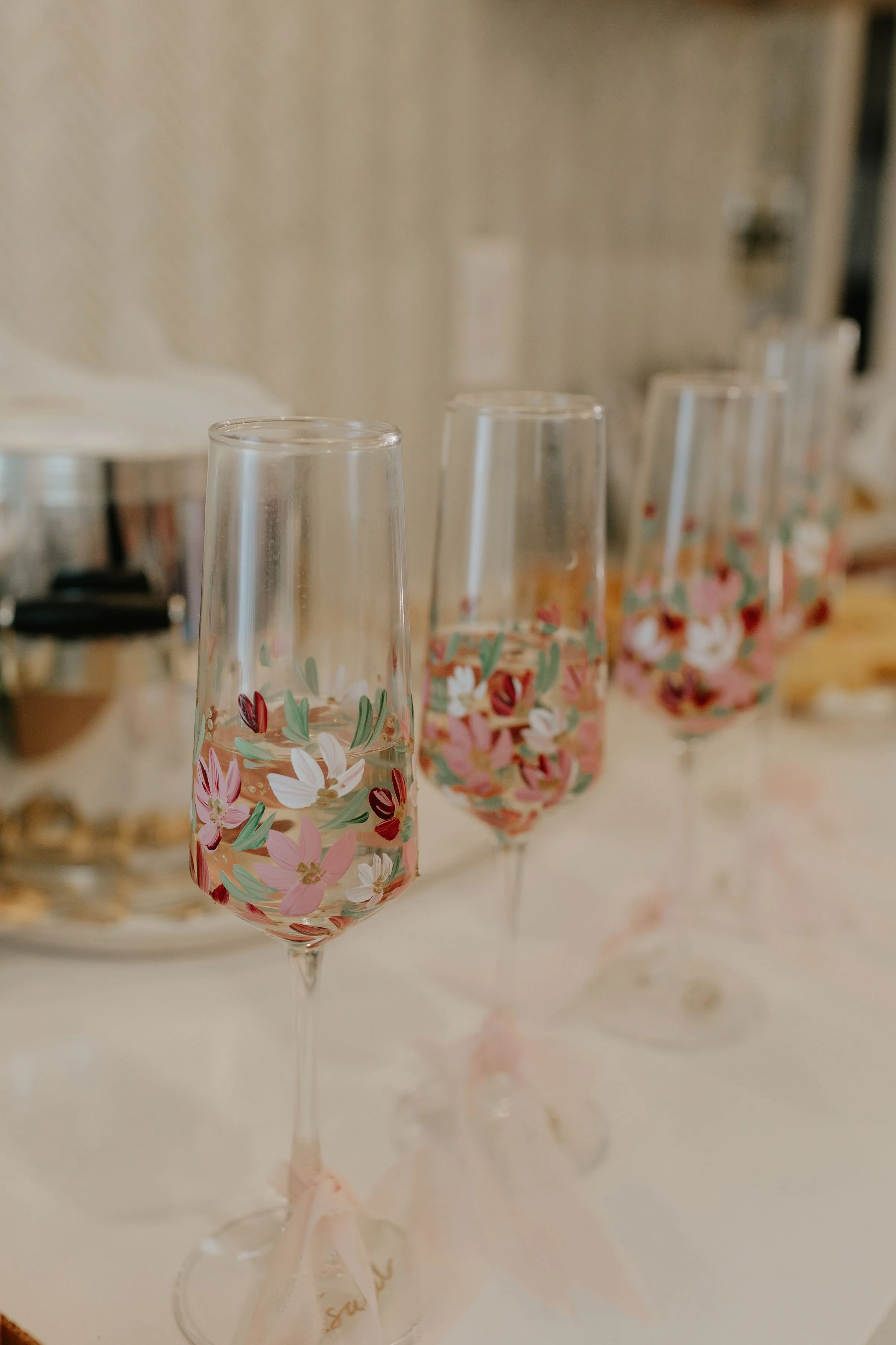 Four champagne flutes with pink, white, and red floral designs filled with pink liquid, placed on a white table.
