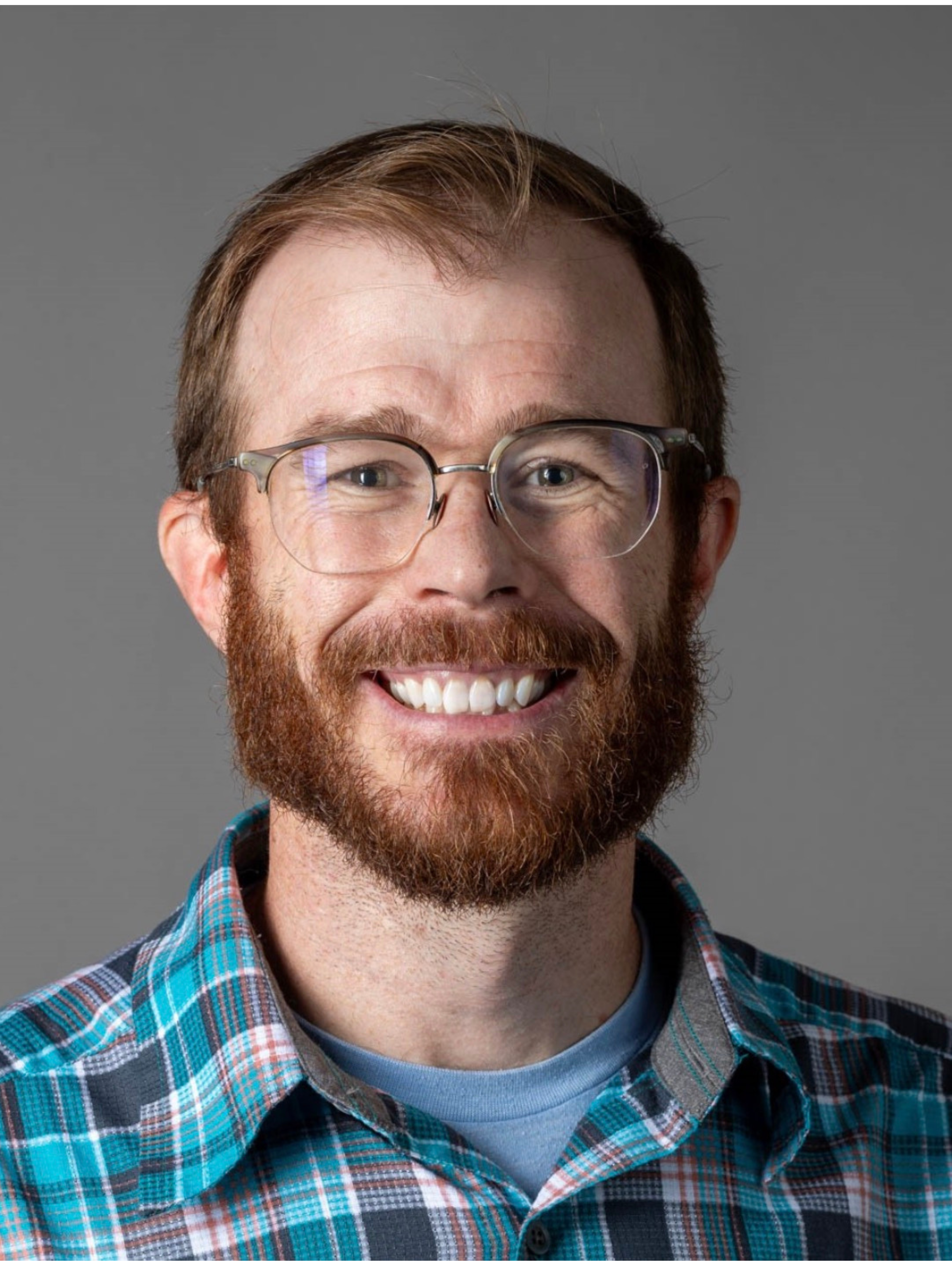 A smiling man with glasses, a red beard, wearing a plaid shirt, against a gray background.