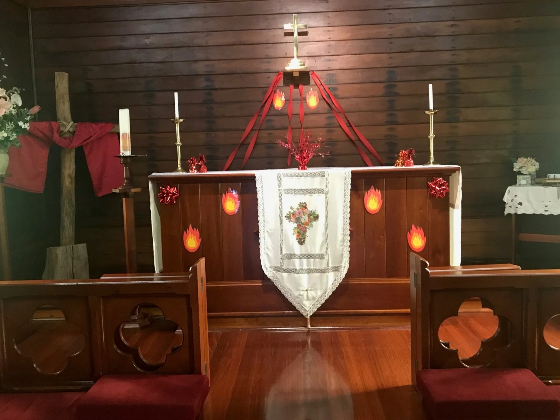 Inside a church altar decorated with a white cloth featuring a floral embroidery, red bows, candles, red ribbons, and paper flames with a cross at the top center.