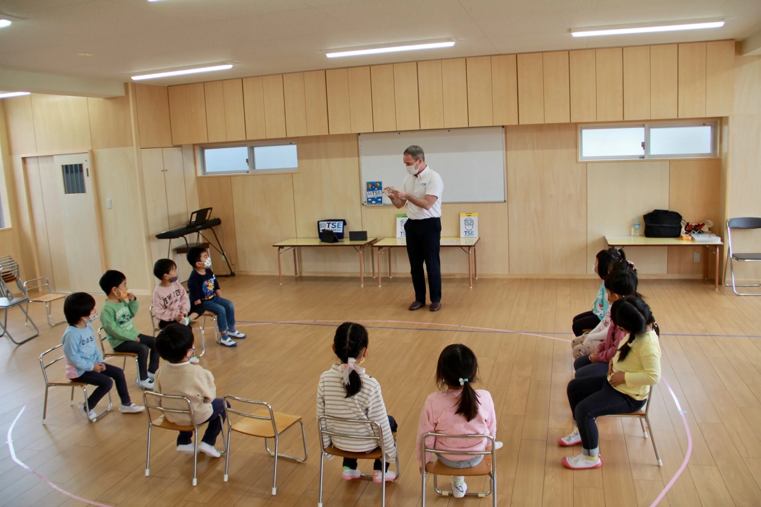 教室で椅子に座っている子供たちと立って話している大人。