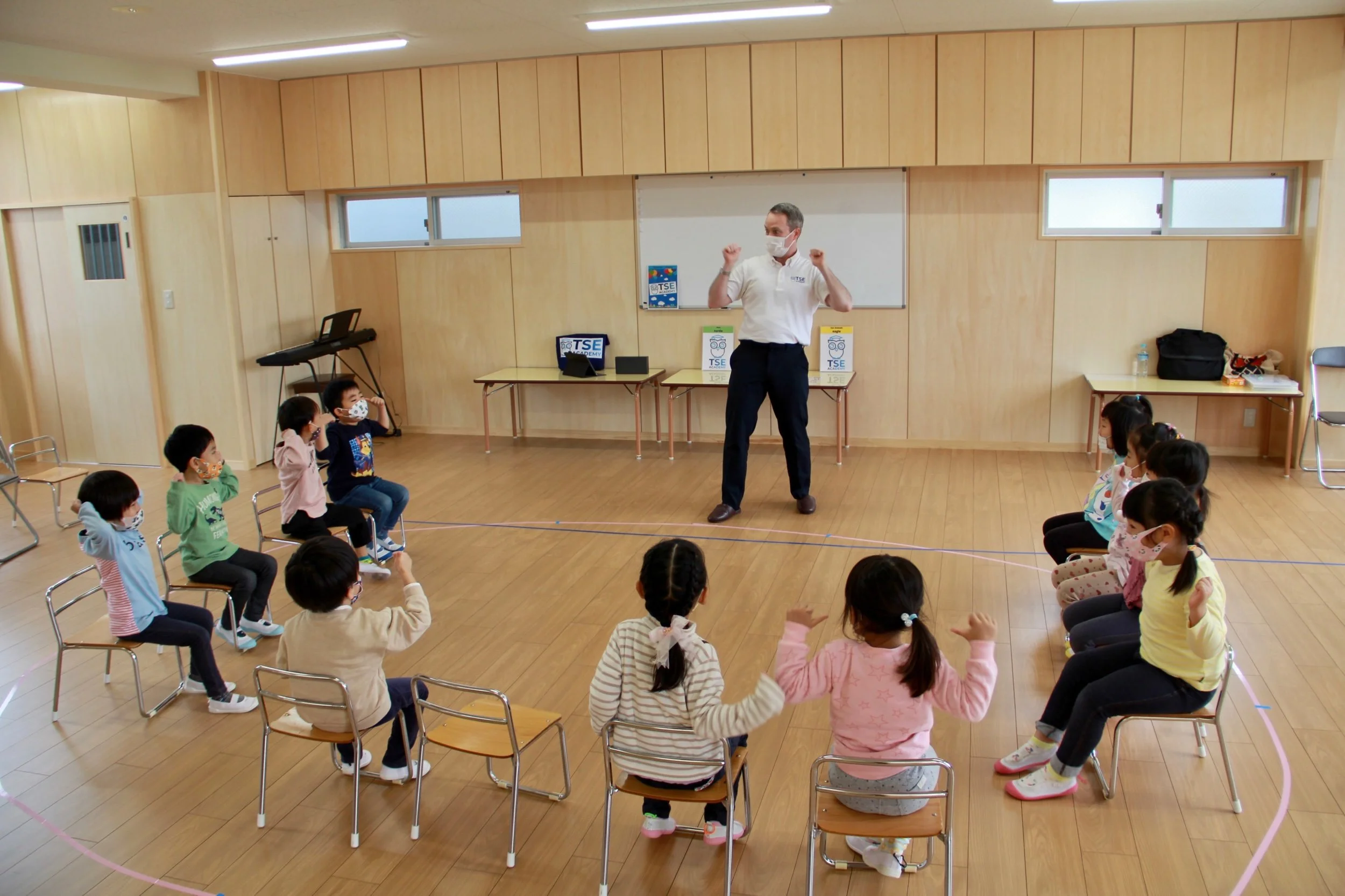 教室で教師が子供たちに指導している様子。子供たちは椅子に座って、マスクを着用し、先生に向かって動作を模倣。部屋は木製の壁と床が特徴的。